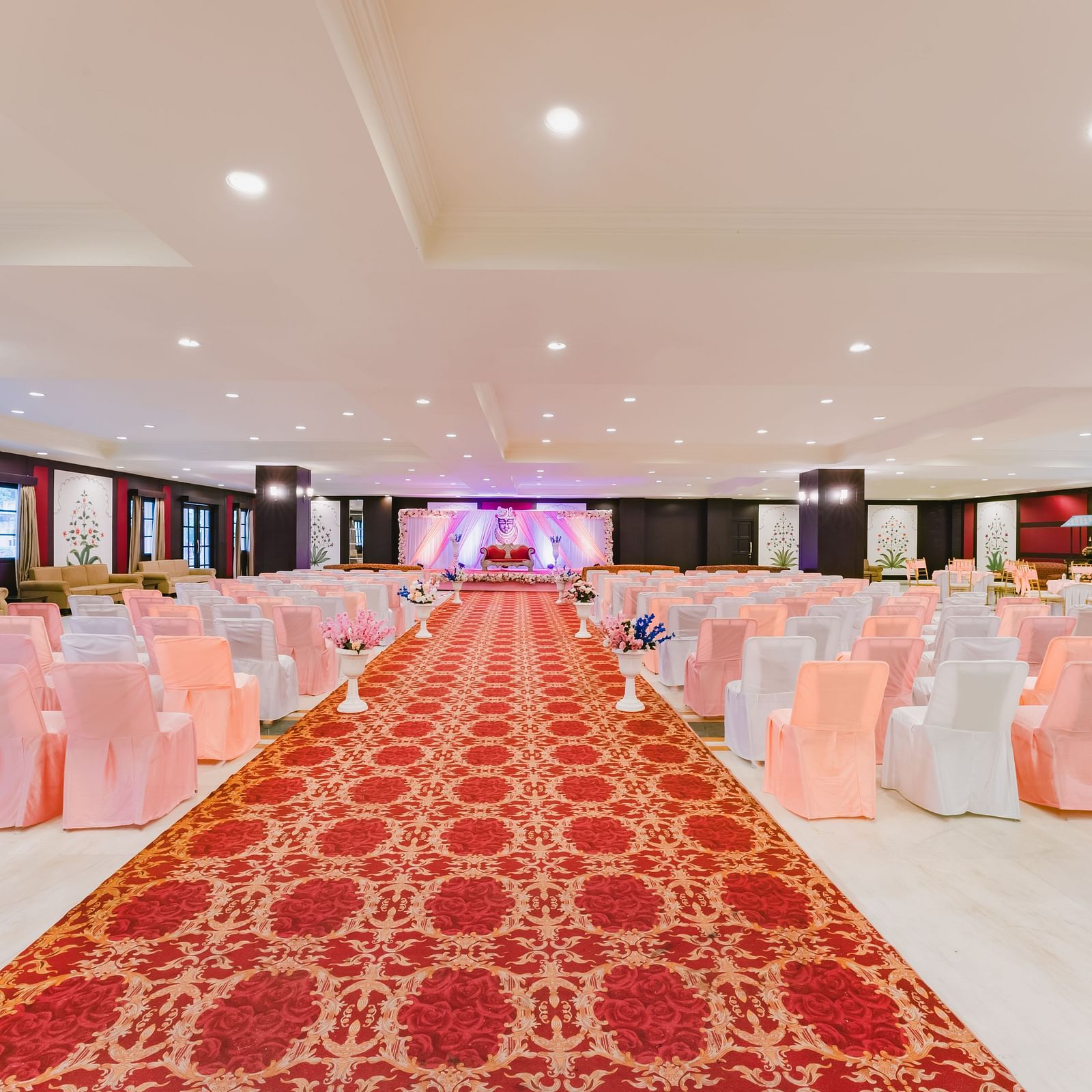 A interior deccorated setup with a red carpet aisle leading to a stage, at the Raj Mahal at Shri Vallabh Vilas Lords Plaza Nathdwara, surrounded by neatly arranged peach chairs.