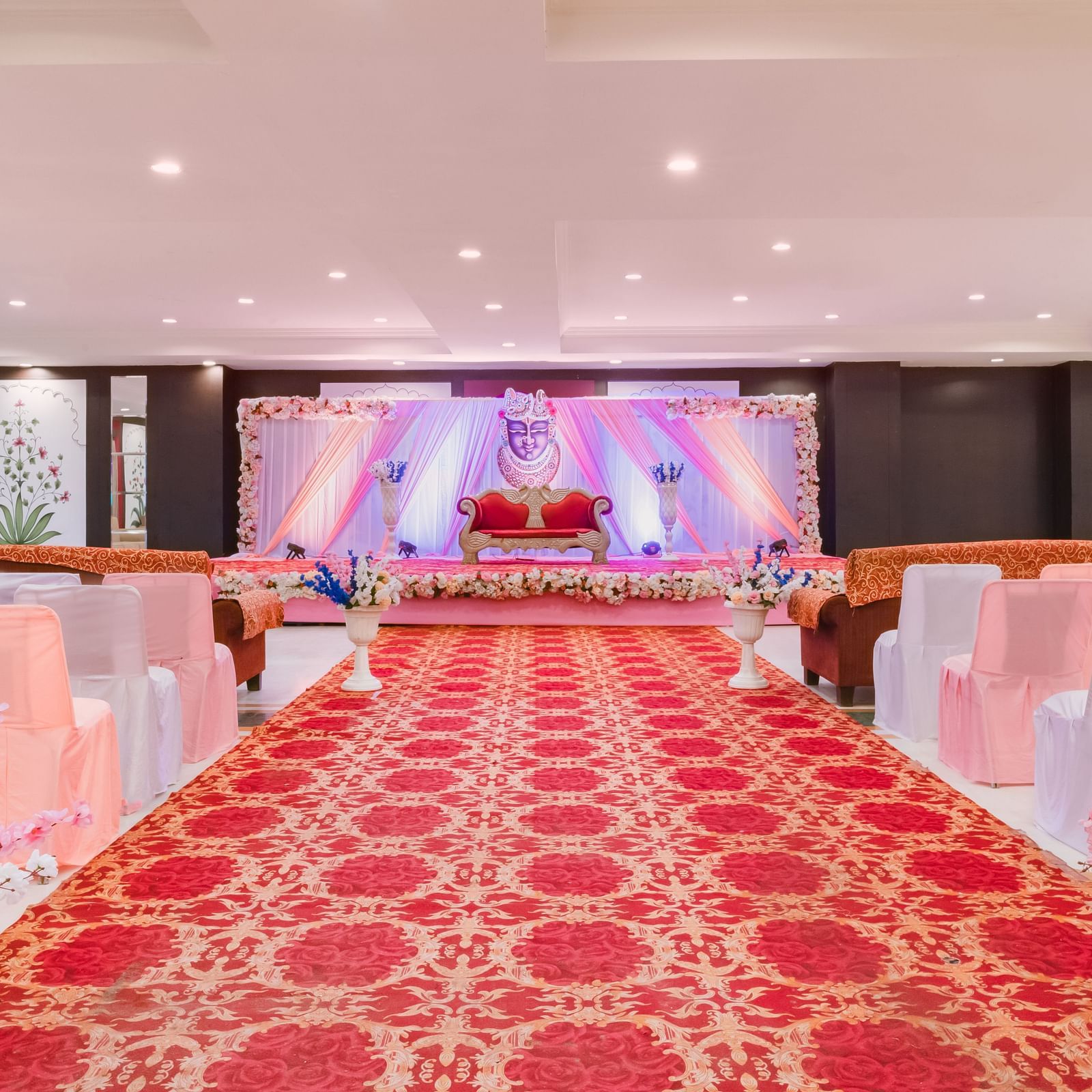 A decorated stage and aisle lined with peach-covered chairs and floral arrangements in Raj Mahal hall at Shri Vallabh Vilas Lords Plaza Nathdwara.