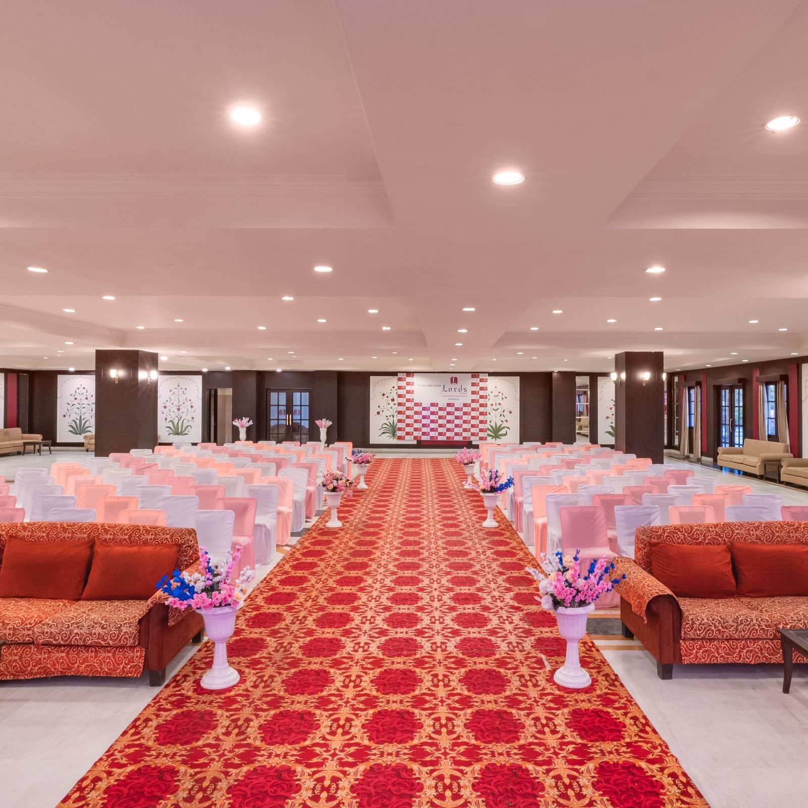 A wide red carpet aisle flanked by peach chairs and sofas in a large indoor Raj Mahal Hall at Shri Vallabh Vilas Lords Plaza Nathdwara.