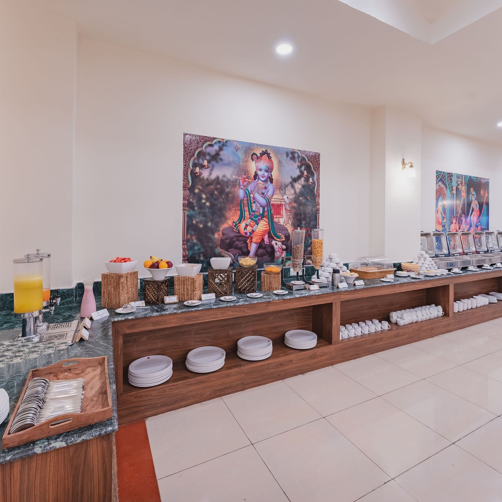 A wider view of the buffet spread with plates, beverages, and artwork on the wall in a well-lit dining area at Shri Vallabh Vilas Lords Plaza Nathdwara.