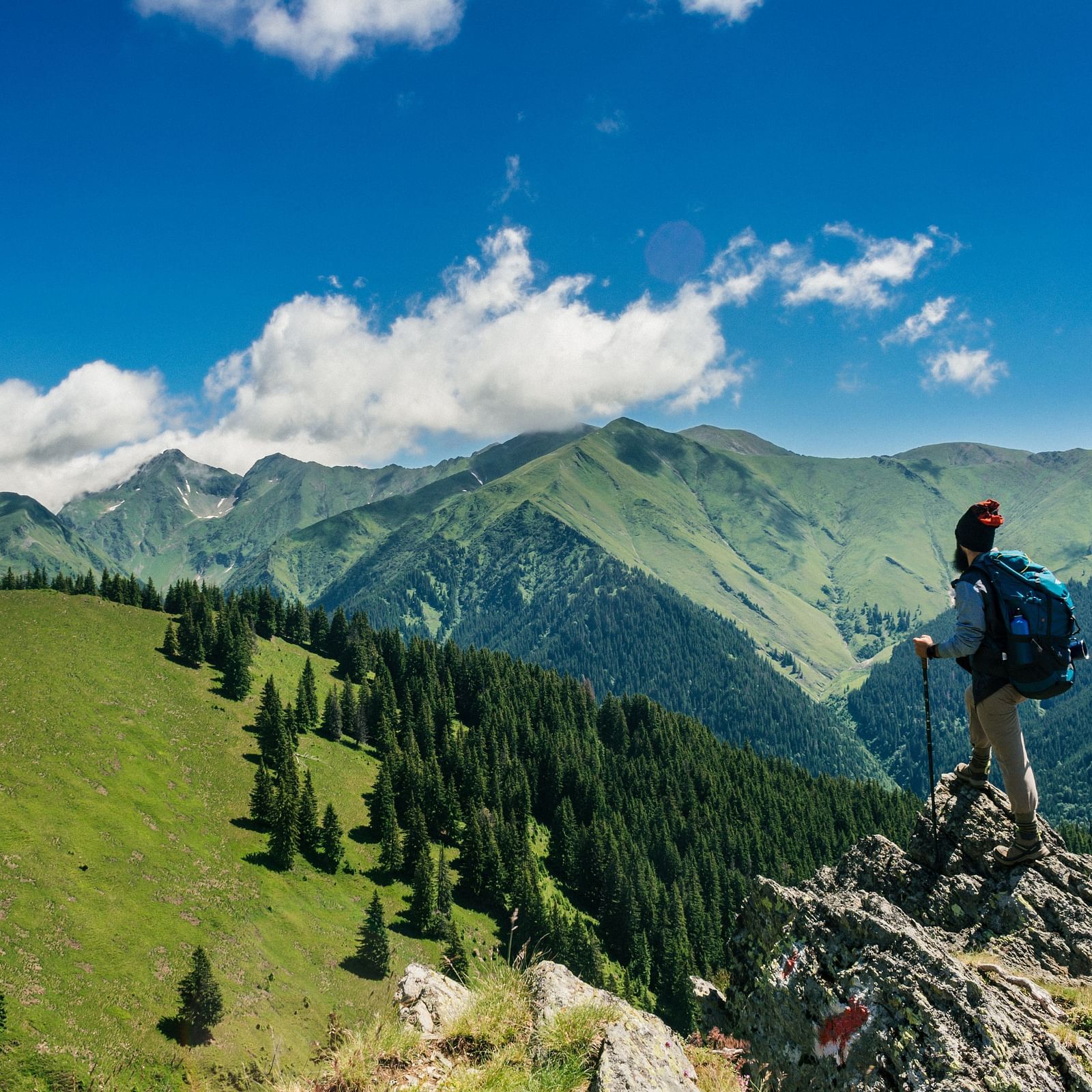A man standing on the edge of a cliff looking at the mountains on a sunny day.