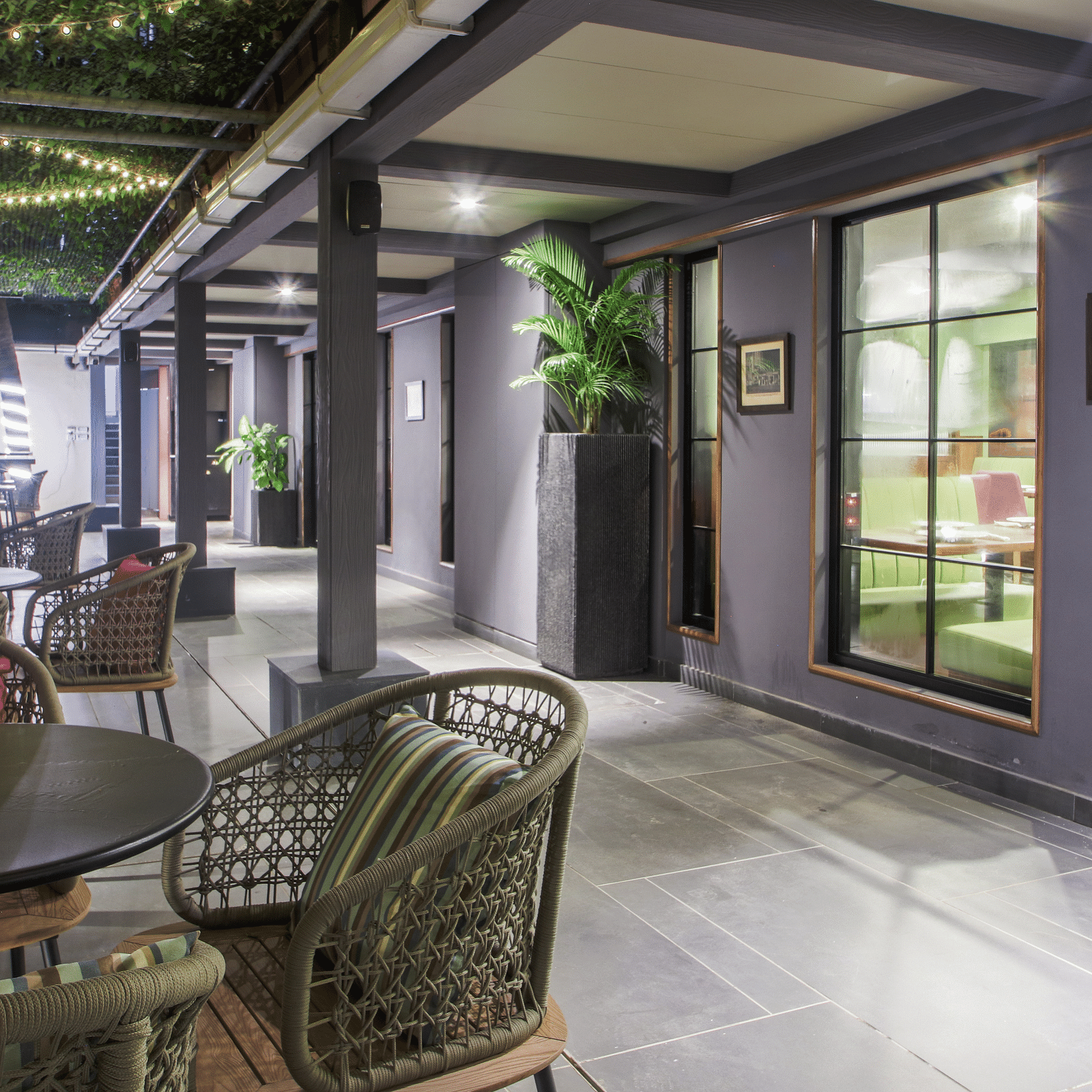 A dimly lit lounge area with several round tables and chairs, as well as a long   | T24 Retronch against the wall. The space is decorated with plants, artwork, and warm lighting, creating a cosy atmosphere.