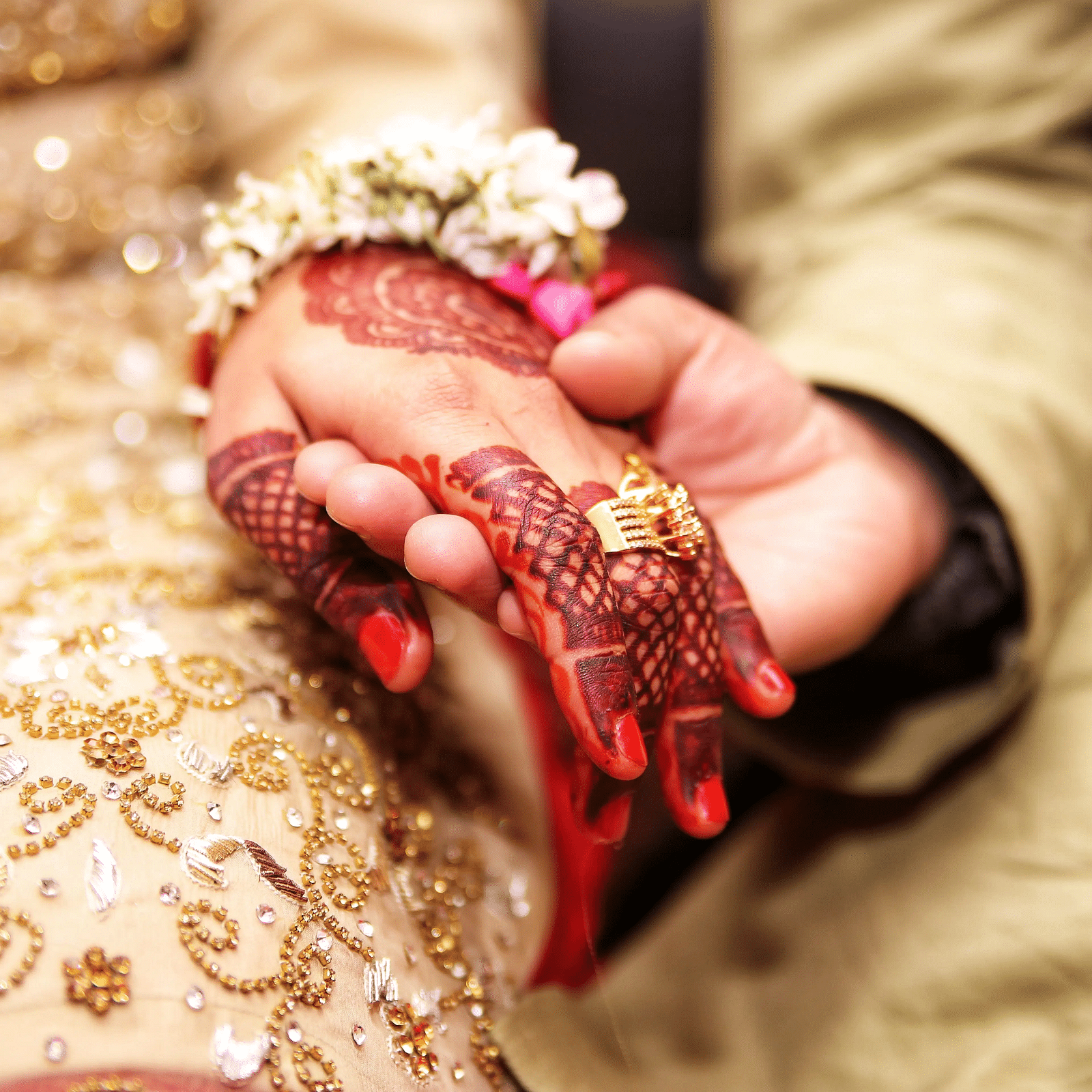 a couple holding hands, with the bride’s hand adorned in intricate henna designs, red bangles, and a floral bracelet