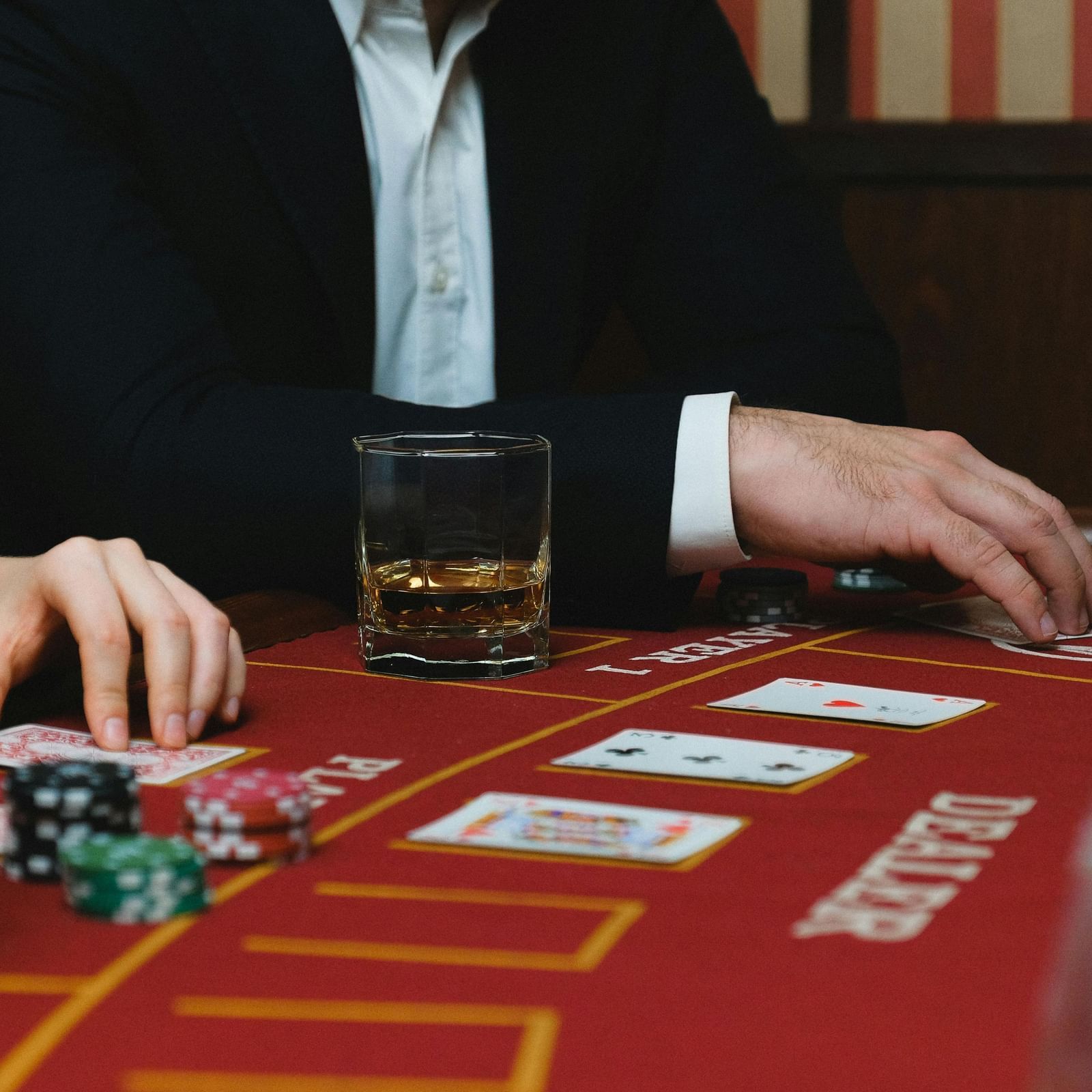 A casino table featuring poker chips and playing cards placed on the table with a whisky glass beside a person's hand.
