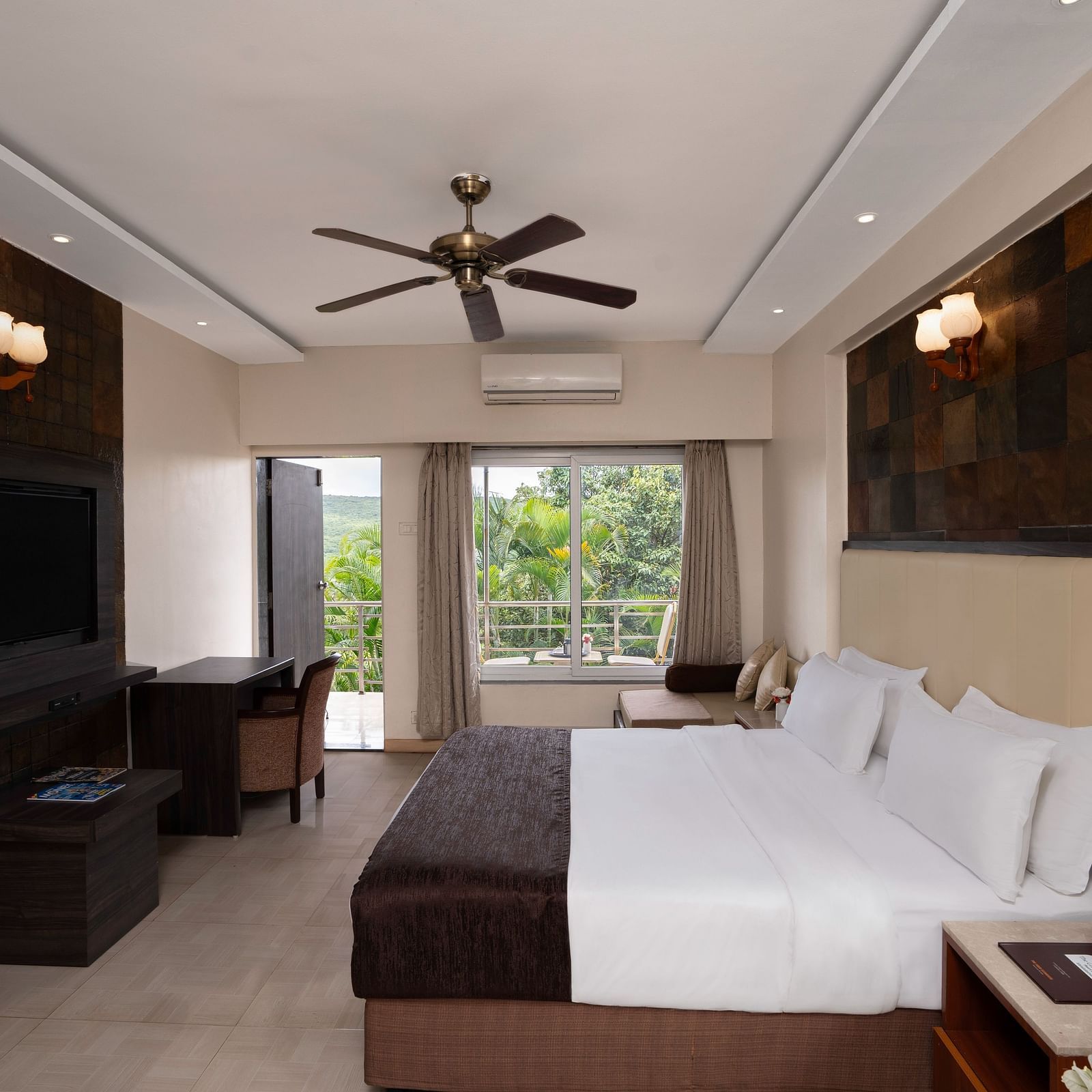 A spacious room with neutral-toned walls, a large bed, a flat-screen TV, and large windows offering natural light.
