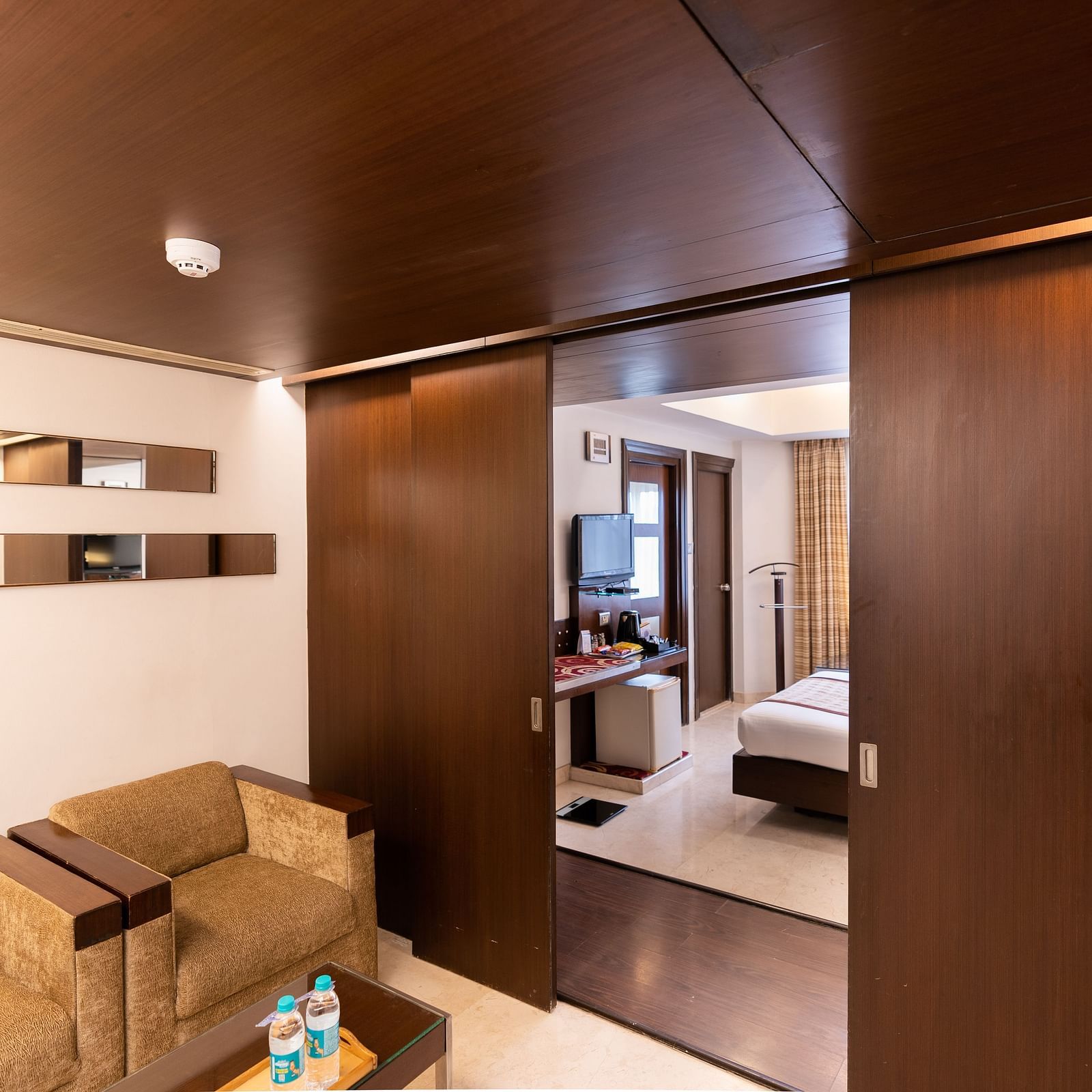 A hotel suite featuring sleek wooden finishes, a cosy sitting area, and a peek into the adjoining bedroom, creating a seamless living space - The President Hotel Jayanagar, Bangalore