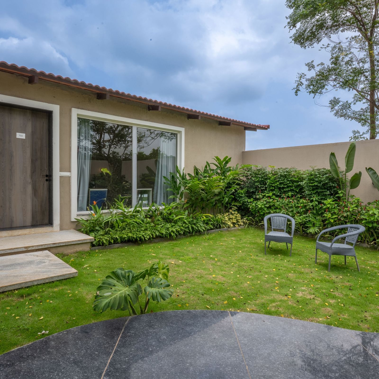 Ananta Garden Villa exterior showing the villa building, a lush green garden with outdoor seating, and cloudy sky.