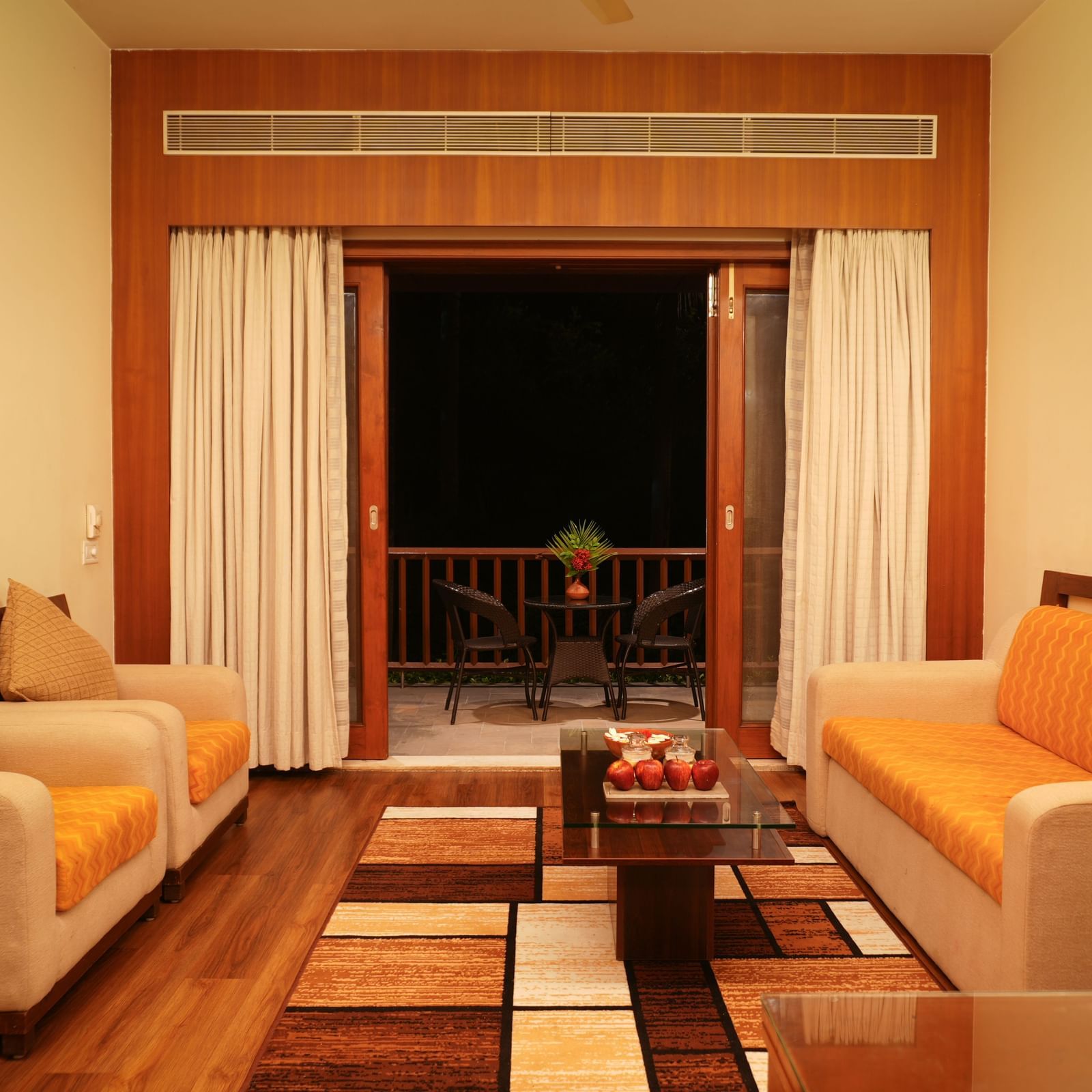 Plush sofas in the living room of the One-Bedroom Suite at Vedic Village Spa Resort, set against carpeted wooden floors and opening to a balcony with a wooden centre table.