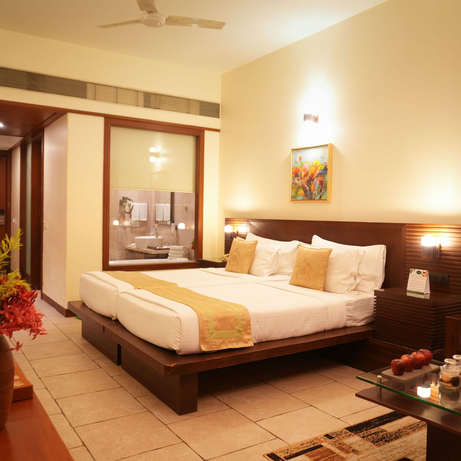 Standard Suite at Vedic Village Spa Resort showcasing a double bed with wooden headrest, tiled flooring, glass-panelled wall, glass-topped table, and a lounge couch.
