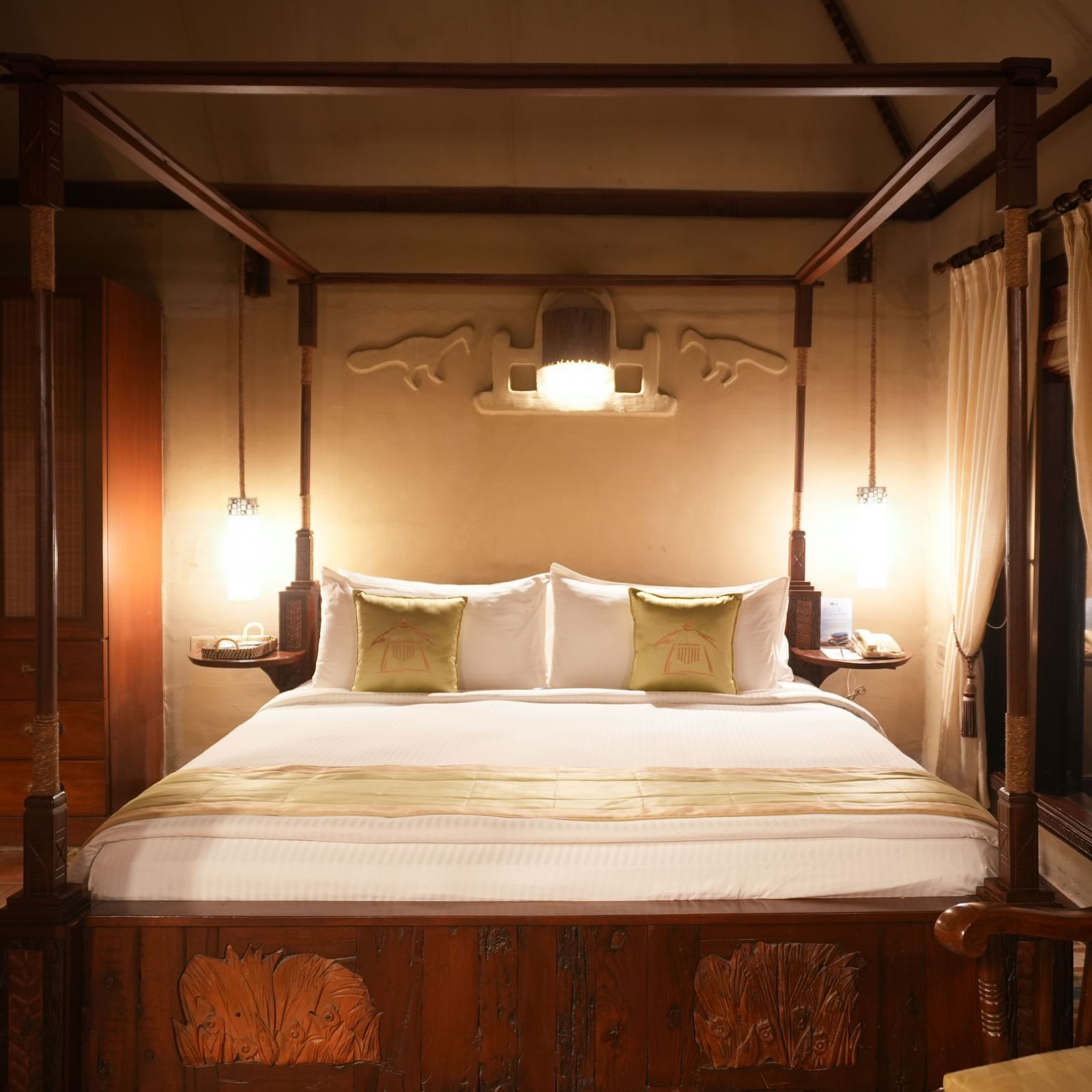 Elegantly styled four-poster king size bed in the Zamindar Suite at Vedic Village Spa Resort, complemented by a wooden lounge chair, and soft lighting.