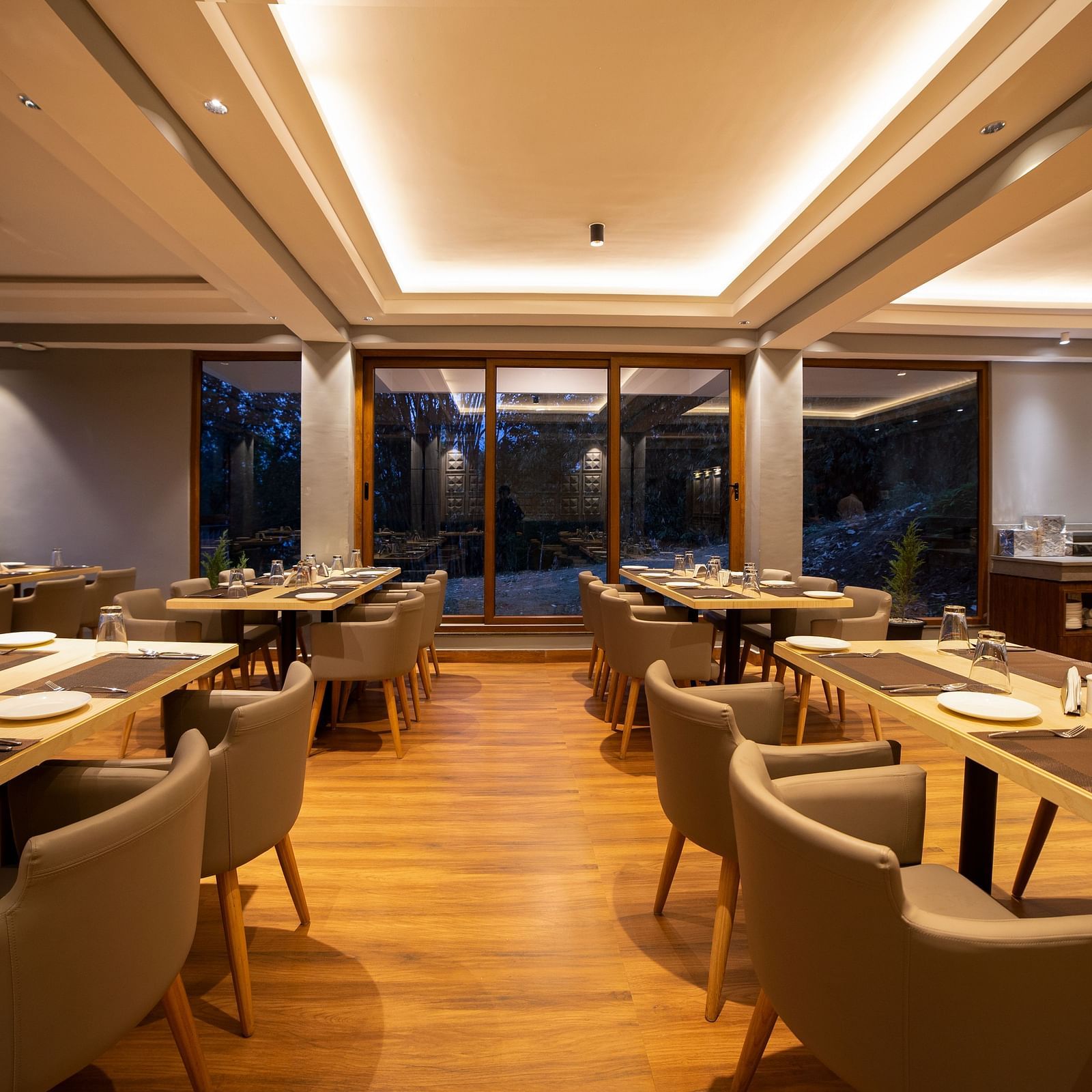 Dining area with wooden flooring, multiple rectangular tables set with plates and glasses, cushioned chairs, recessed ceiling lighting, and large glass windows and doors overlooking trees outside - Yashshree Pelling Chekhim Retreat.