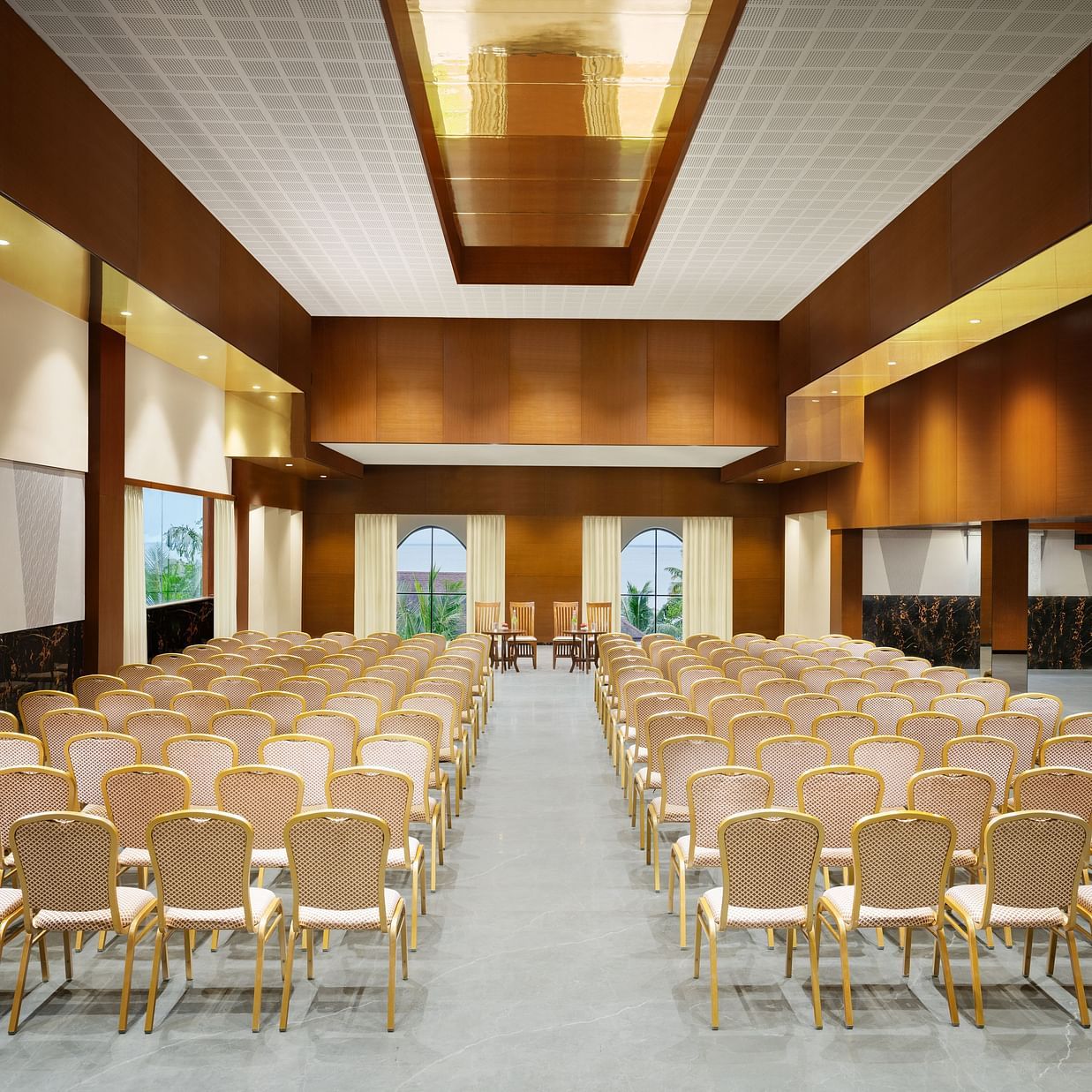 A well-lit event hall with chairs arranged neatly facing a central stage, featuring modern wooden interiors, windows offering natural light, and a sleek, polished floor - Uday Backwater Resort