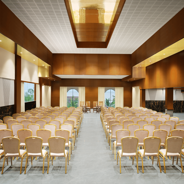 A well-lit event hall with chairs arranged neatly facing a central stage, featuring modern wooden interiors, windows offering natural light, and a sleek, polished floor - Uday Backwater Resort