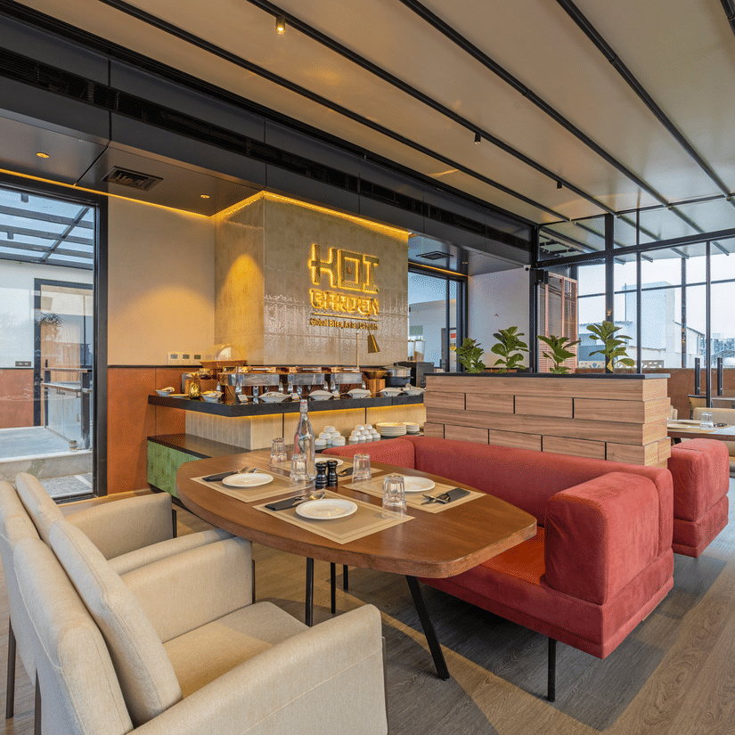 Indoor dining area at Koi Garden restaurant within Hotel Bawa Gurgaon. Tables and chairs are arranged across the room with a buffet counter in the background. Glass walls line the side of the space.