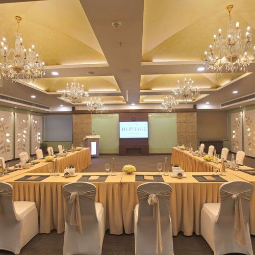 Well-lit conference room with a U-shaped table, elegant chandeliers, and a screen displaying HERITAGE - Heritage Village Resorts & Spa.