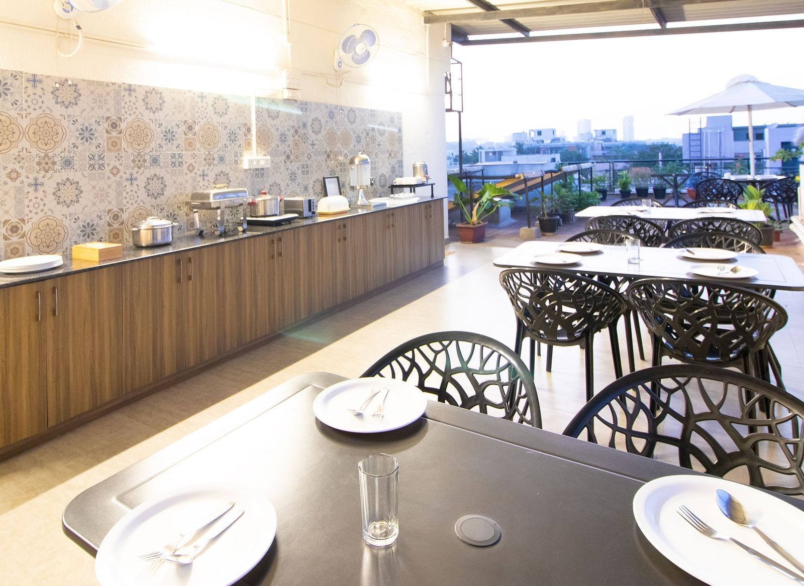 A dining space with tables and chairs under a partial roof, featuring a buffet setup along one side at Sanctum Suites Indiranagar, Bangalore.
