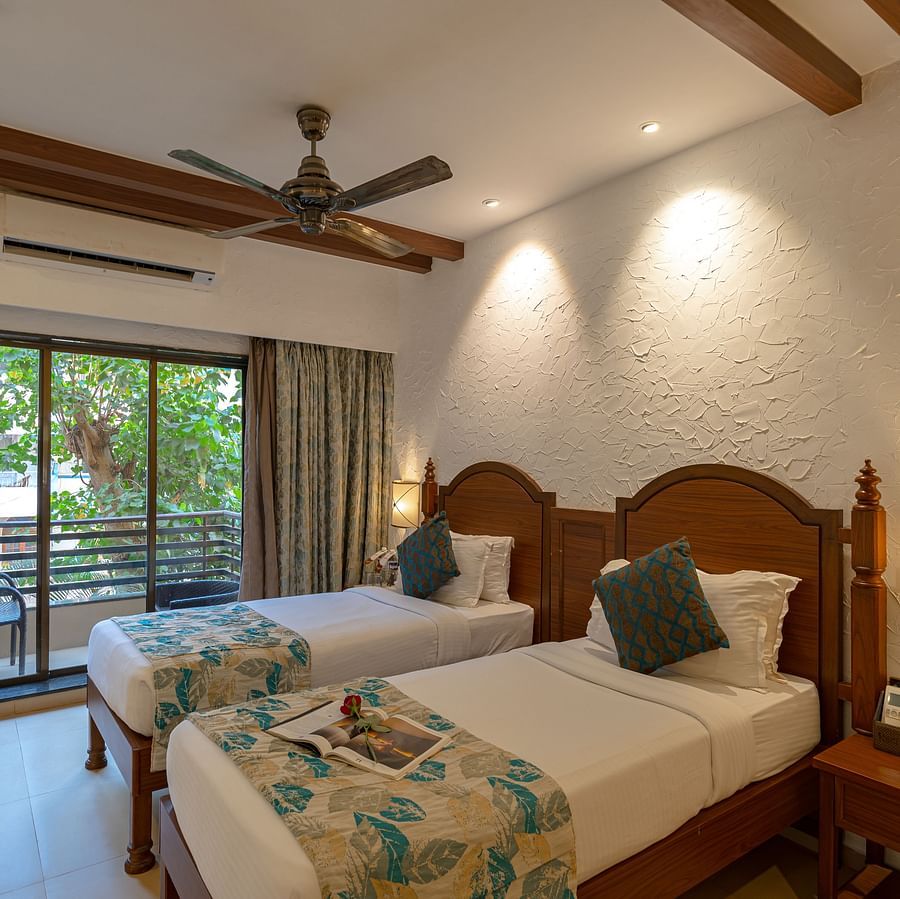 An image of a hotel room with twin beds dressed in white sheets, a cosy throw, wall-mounted lamps on each side, and an attached balcony.