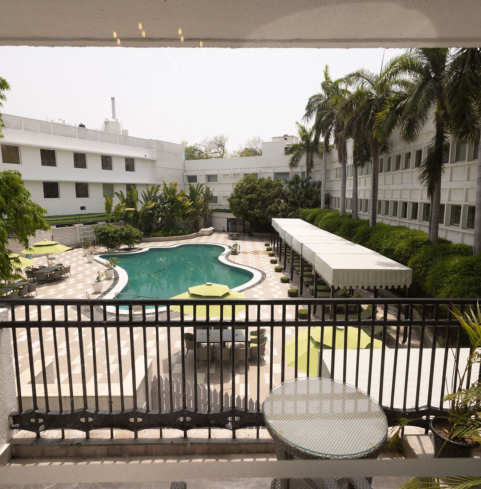 Luxury King Bed Room with Balcony at The Claridges New Delhi, offering a view of the hotel’s outdoor open-air swimming pool surrounded by lush trees and nearby buildings