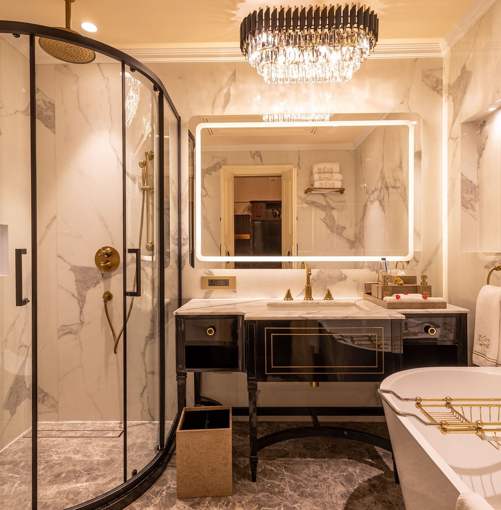 Executive Suite washroom at The Claridges New Delhi, featuring a sleek sink, wall-mounted mirror, glass-enclosed bath area, and a chandelier