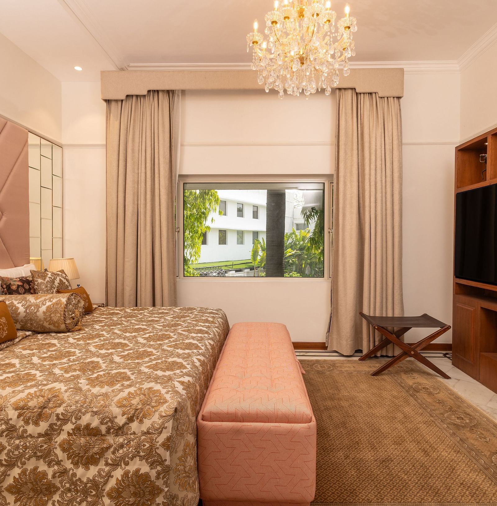 Luxury Suite at The Claridges New Delhi, featuring a cosy bed, wall-mounted TV, chandelier overhead, and a serene view through the window