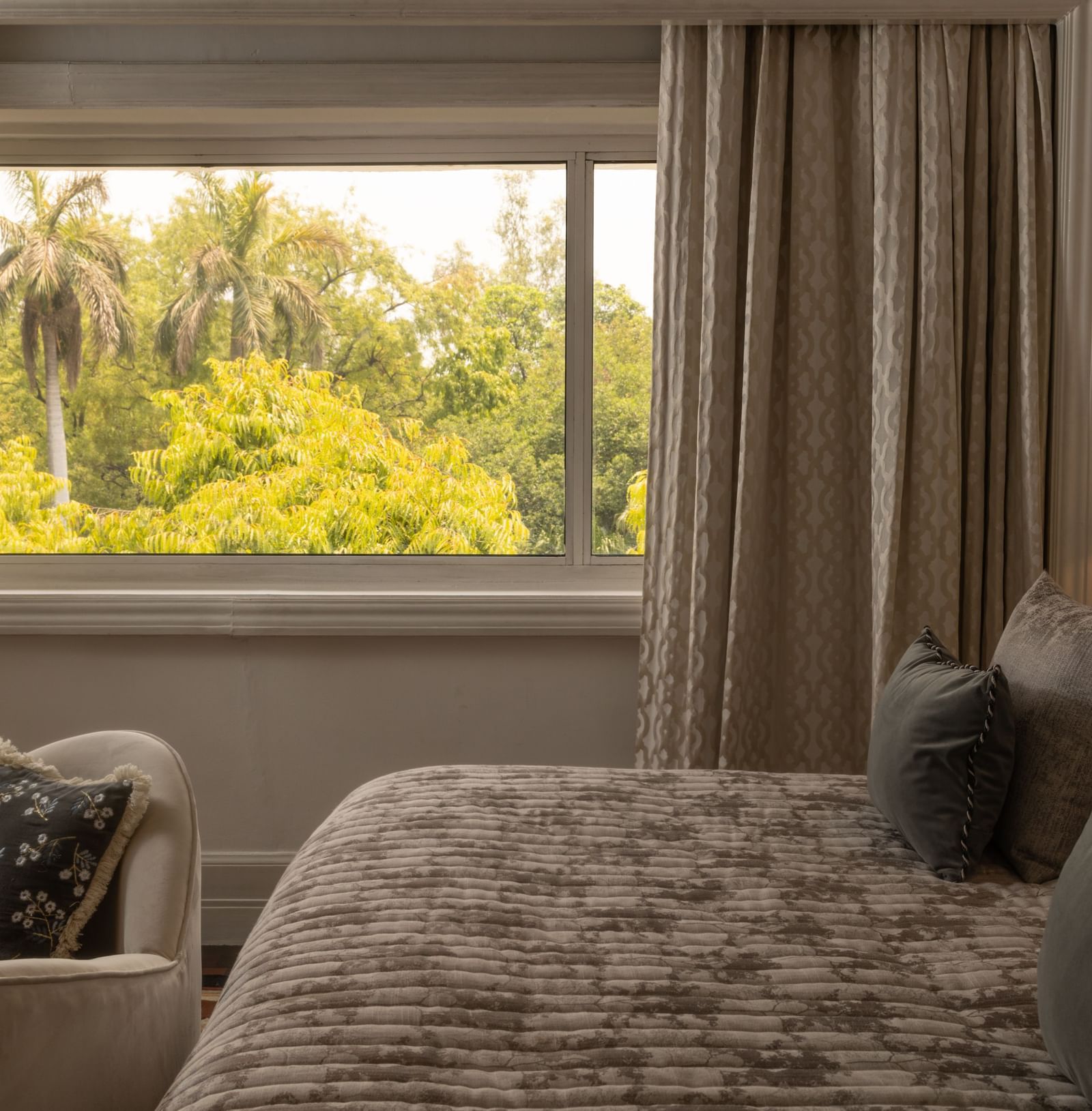 The Claridges New Delhi guest room has a patterned bedspread, dark curtains, and a view of palm trees outside.