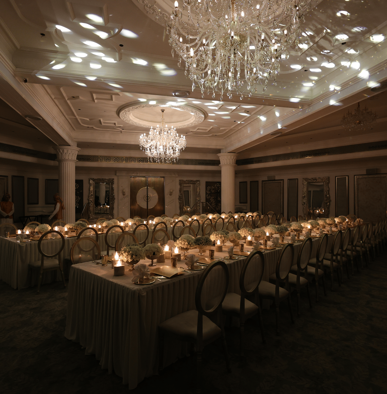 Dimly lit luxury banquet hall featuring a long dining table setup with candles and decorative ceiling light projections under chandeliers.