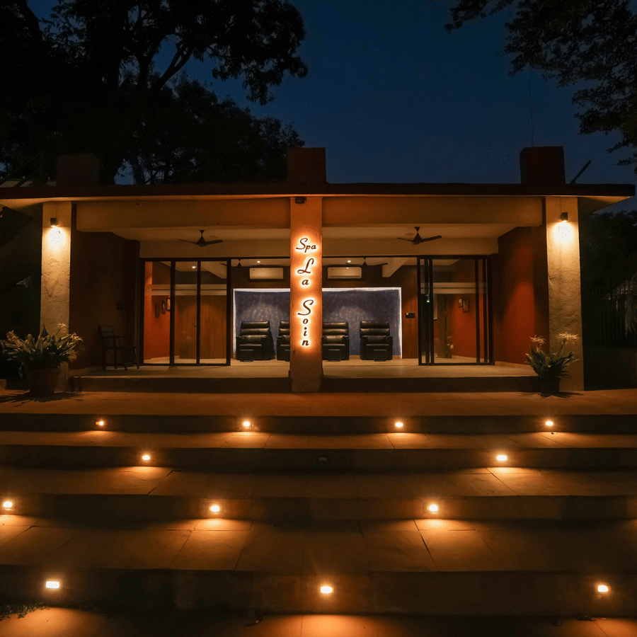Exterior night view of the main building at Adamo The Village showing a modern structure with floor-to-ceiling glass doors and series of steps leading up to the entrance lined on both sides by small ground lights.