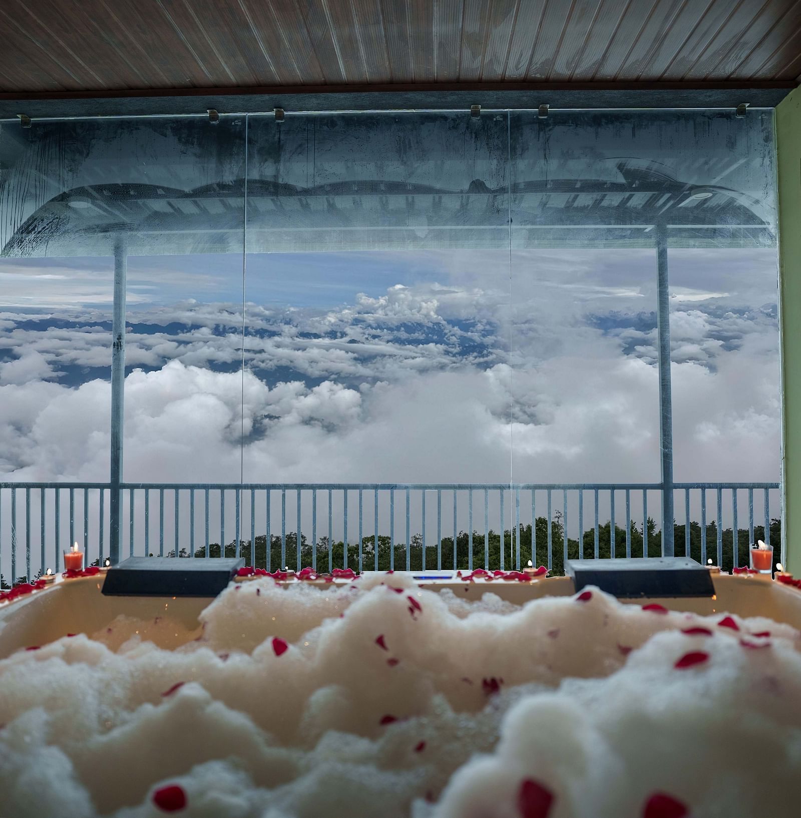 A jacuzzi with rose petals scattered, leading to a balcony overlooking a cloudy sky and distant hills, suggesting a romantic setup at Summit Enigma Resort and Spa.