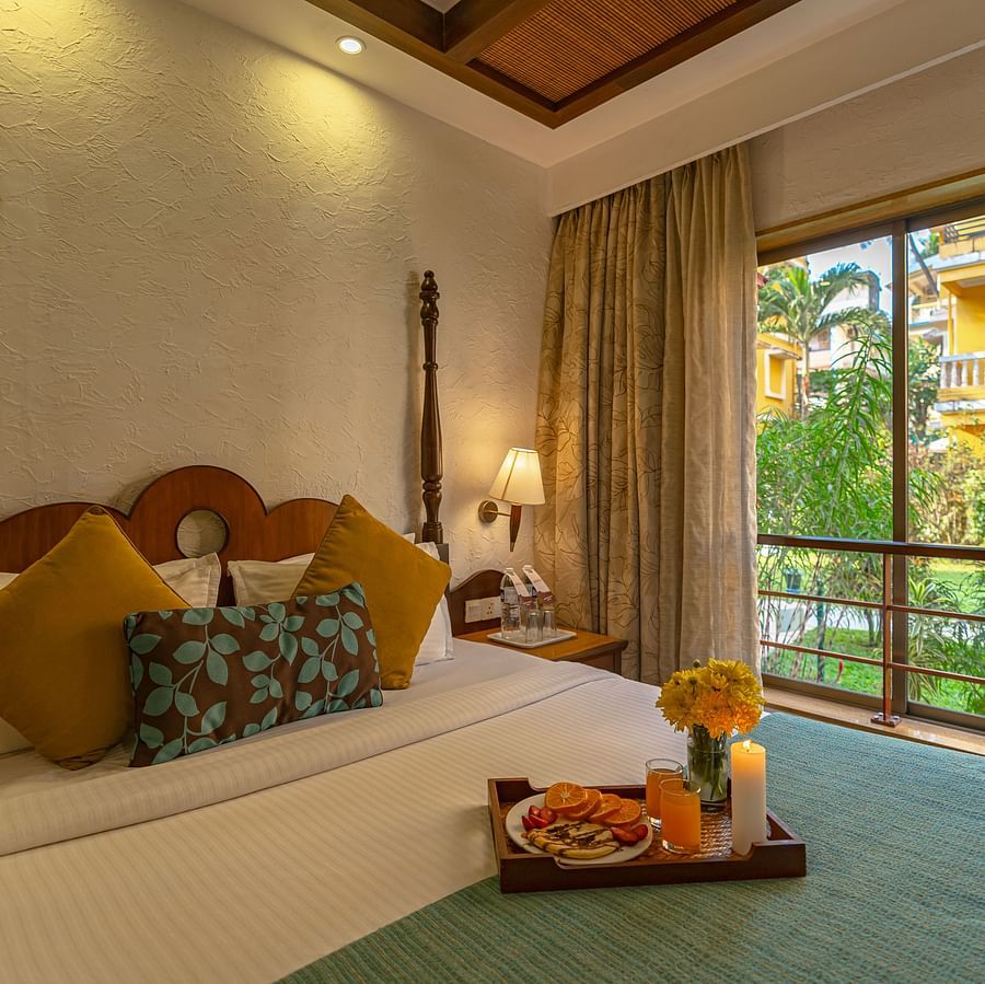 An image of a room showcasing a beautifully decorated bed adorned with a tray of candles, flowers, and fruits, complemented by a large window.