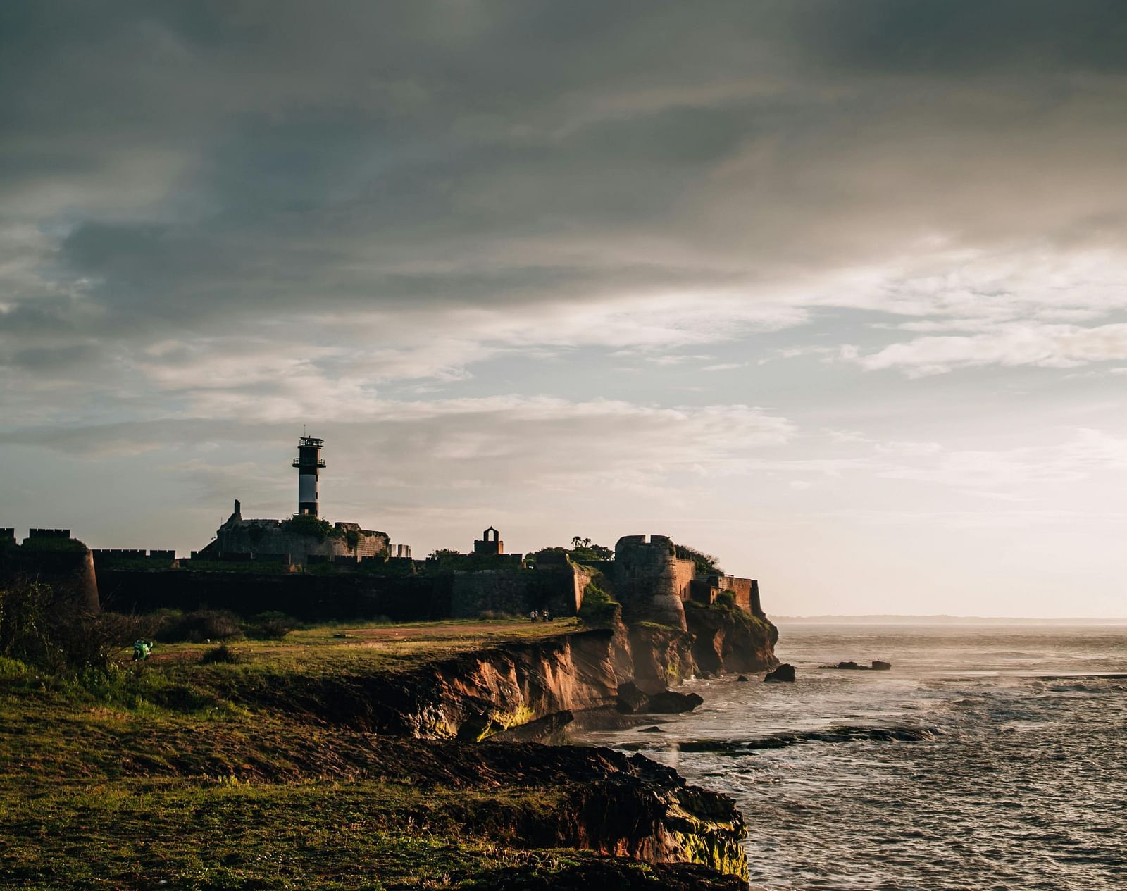 A wide angle shot featuring a cliffside overlooking a vast ocean with a cloudy sky in the background.