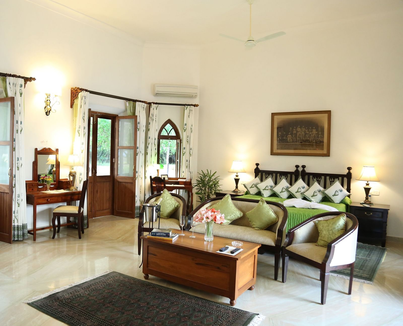 A well-lit room featuring a seating area with a sofa and chairs, a bed with decorative pillows, and multiple doors leading to an outdoor space- Shahpura Bagh.