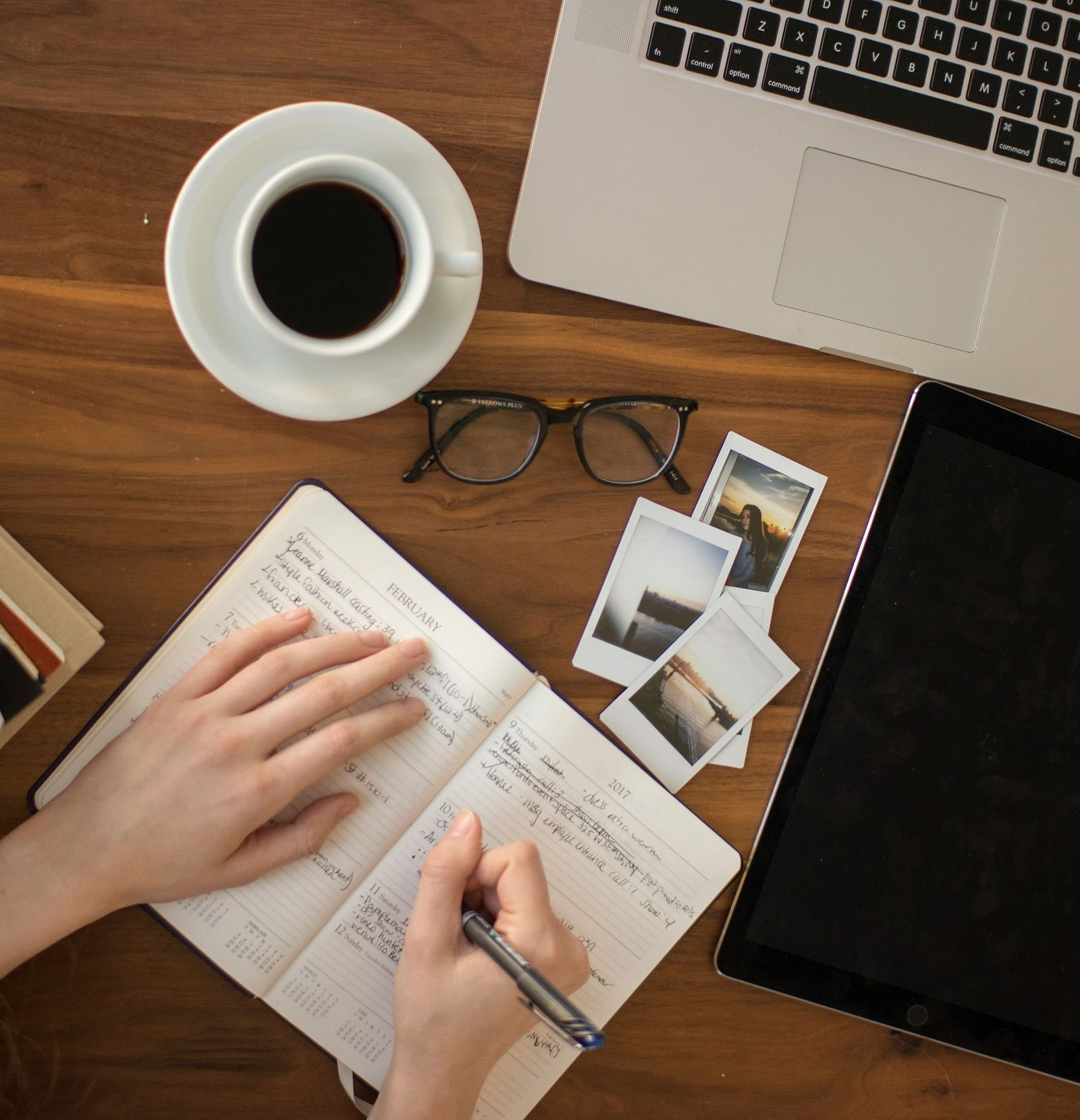 A person writing in a book with a laptop, a cup of coffee and glasses on the table