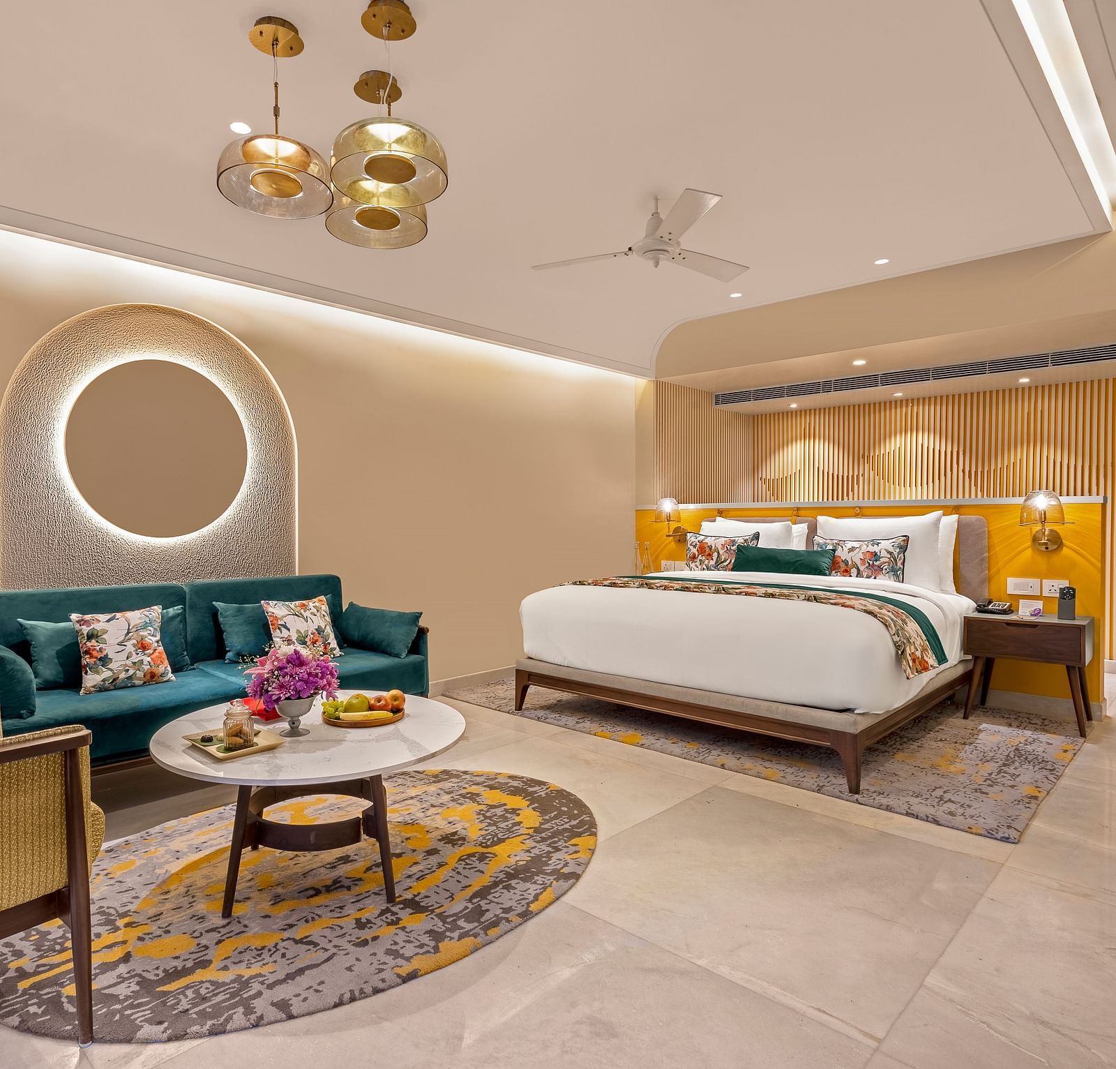 An image of a room featuring a king-size bed, a comfortable sofa, a coffee table, and a television. The room is decorated with a circular mirror, a ceiling fan, and stylish lighting fixtures. 