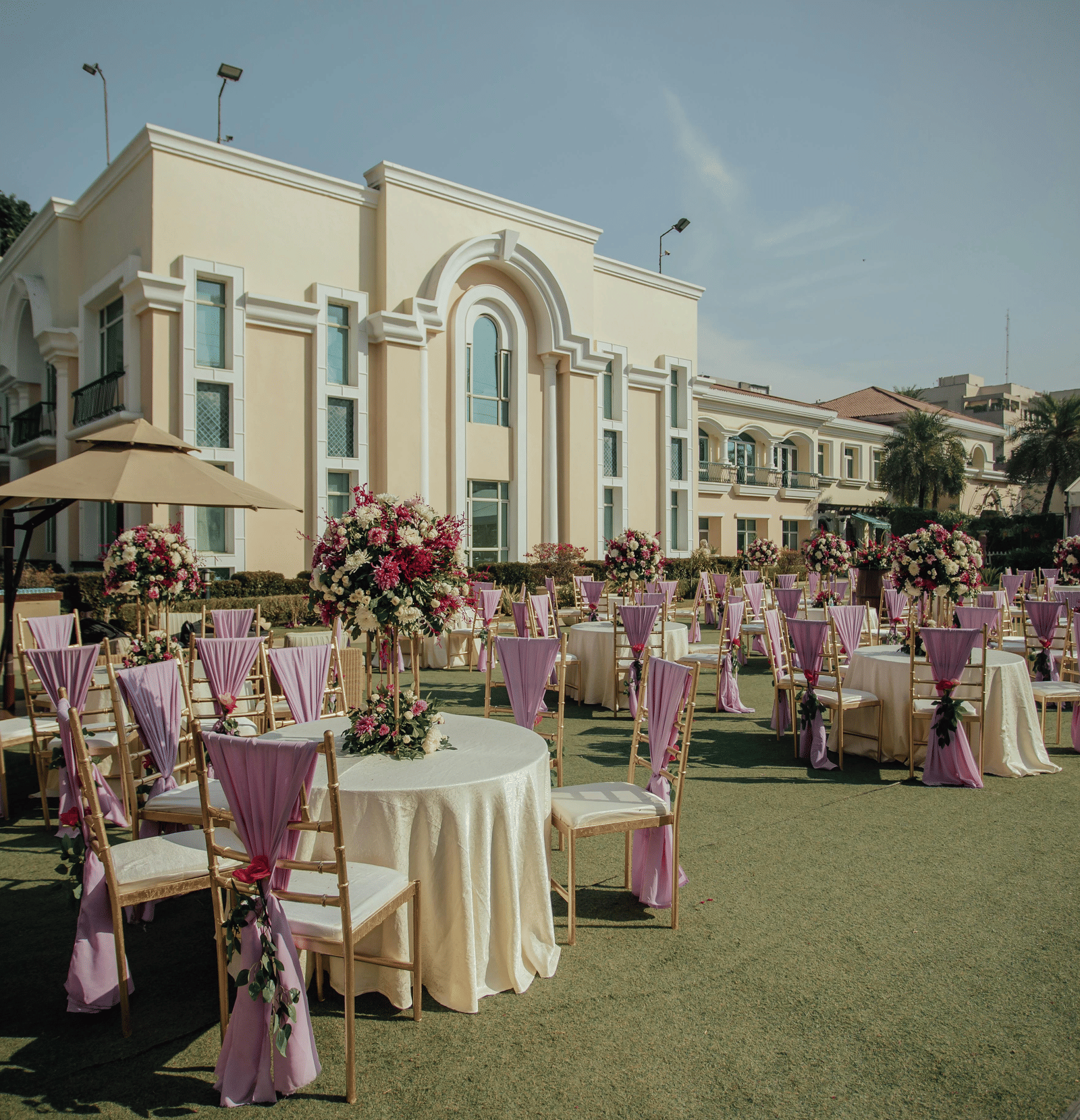 An outdoor wedding setup with round tables, floral decor, and a building in the background.
