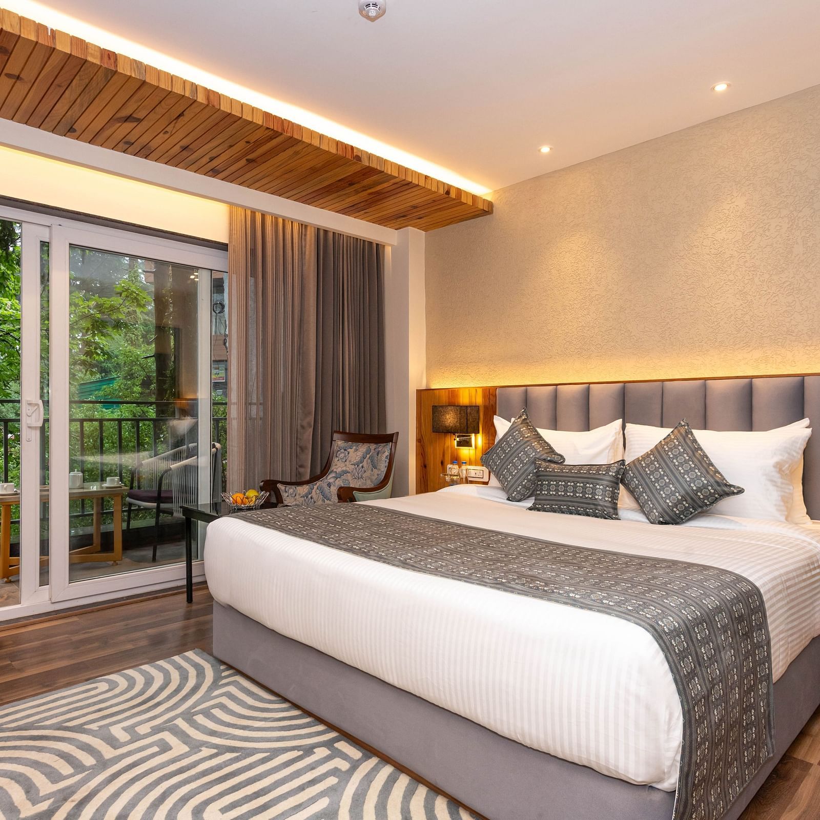 An image of a Superior Room with bed, bed side table, lamps, rug, and an expansive window leading to the balcony - The Belvedere Himalayan Retreat, McLeodganj