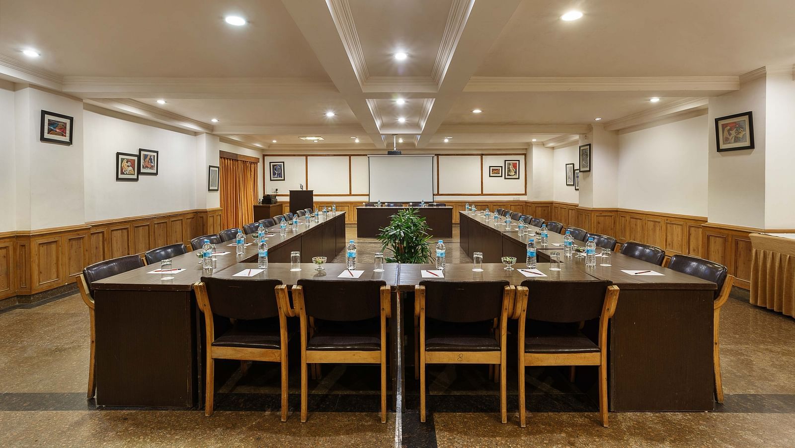 A spacious banquet hall features chairs arranged in a U-shape, illuminated by ceiling lights, and adorned with small photo frames on the walls