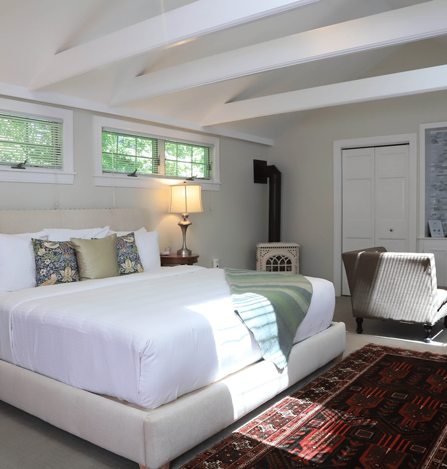 A wide view of a guest room with a large bed, chair, lamp, and wood burning stove.