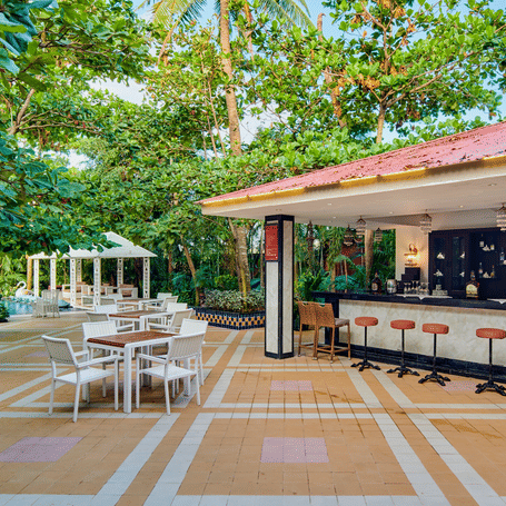 A pool bar area outside at MAYFAIR Palm Beach Resort, Gopalpur-on-Sea. Stools stand along a counter under a roof structure. Tables and chairs occupy a patio. Trees and plants surround the location.