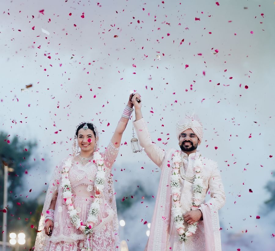 Bride and groom raising their hands as rose petals shower down during a celebration outdoors.
