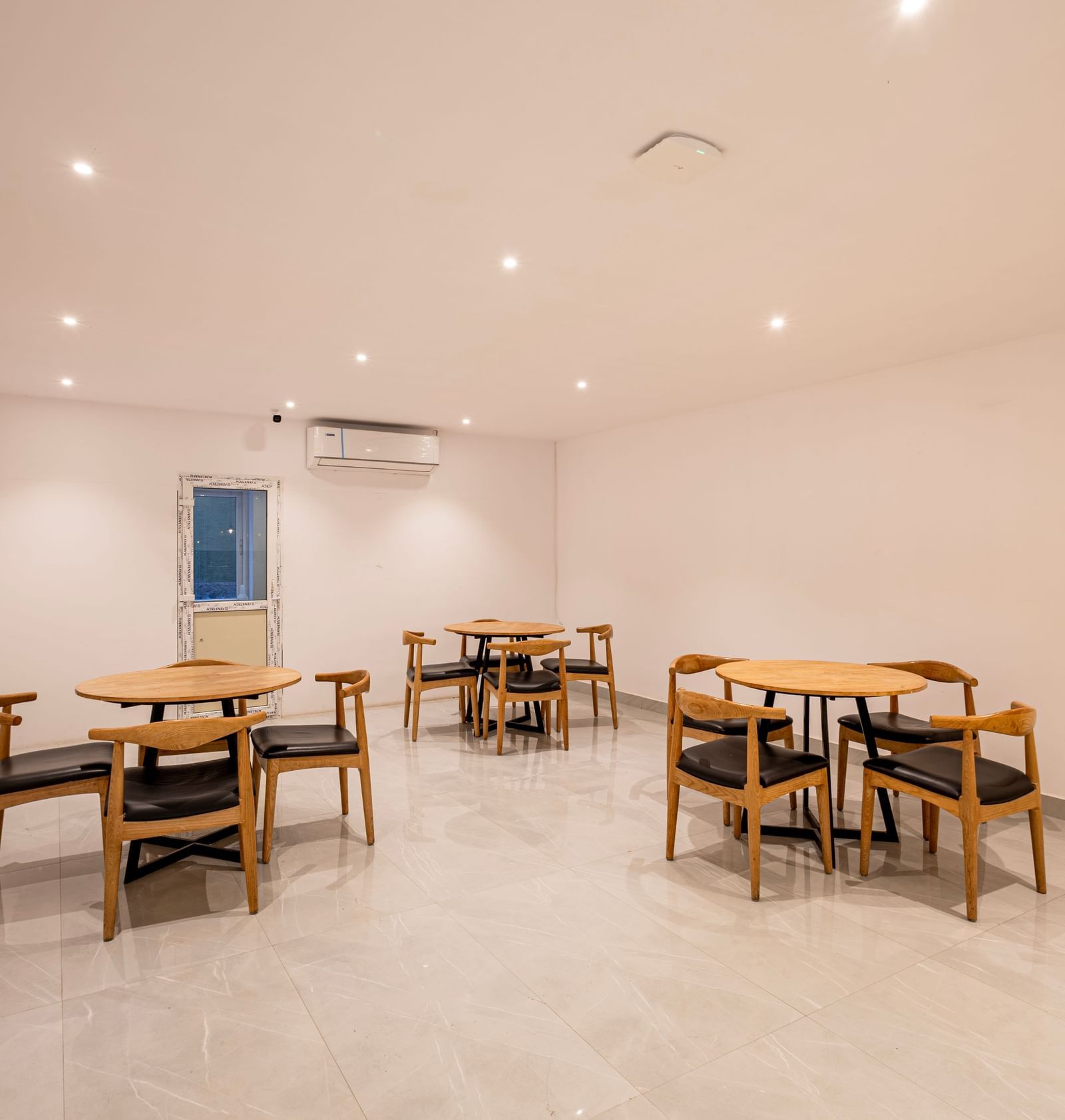 A brightly lit, minimalist dining or common area with several wooden tables and chairs arranged on a light-colored floor - Beestay Hotels Financial District, Hyderabad 
