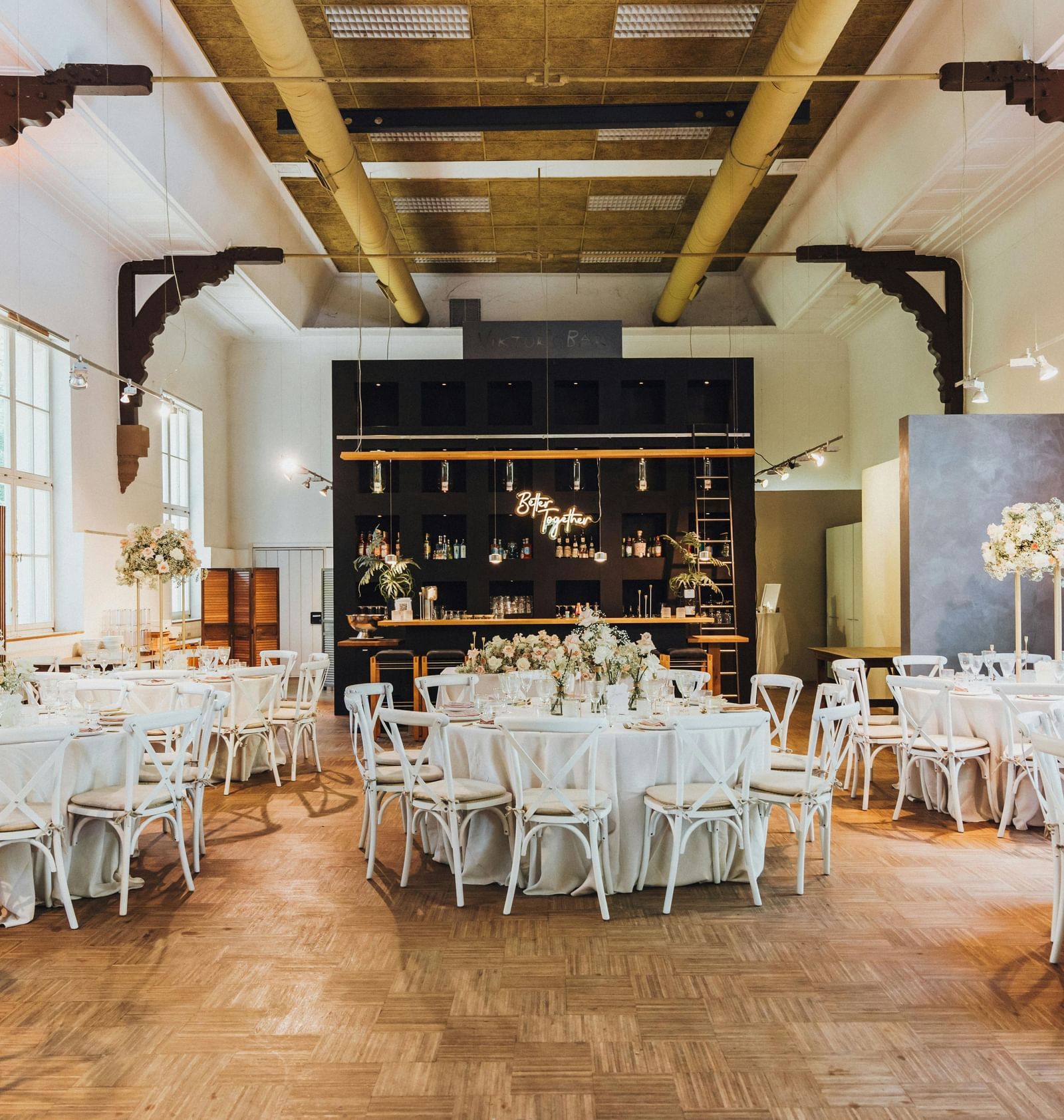 A large, elegant event space with round tables, white chairs, floral centerpieces, and a modern bar with a neon sign.