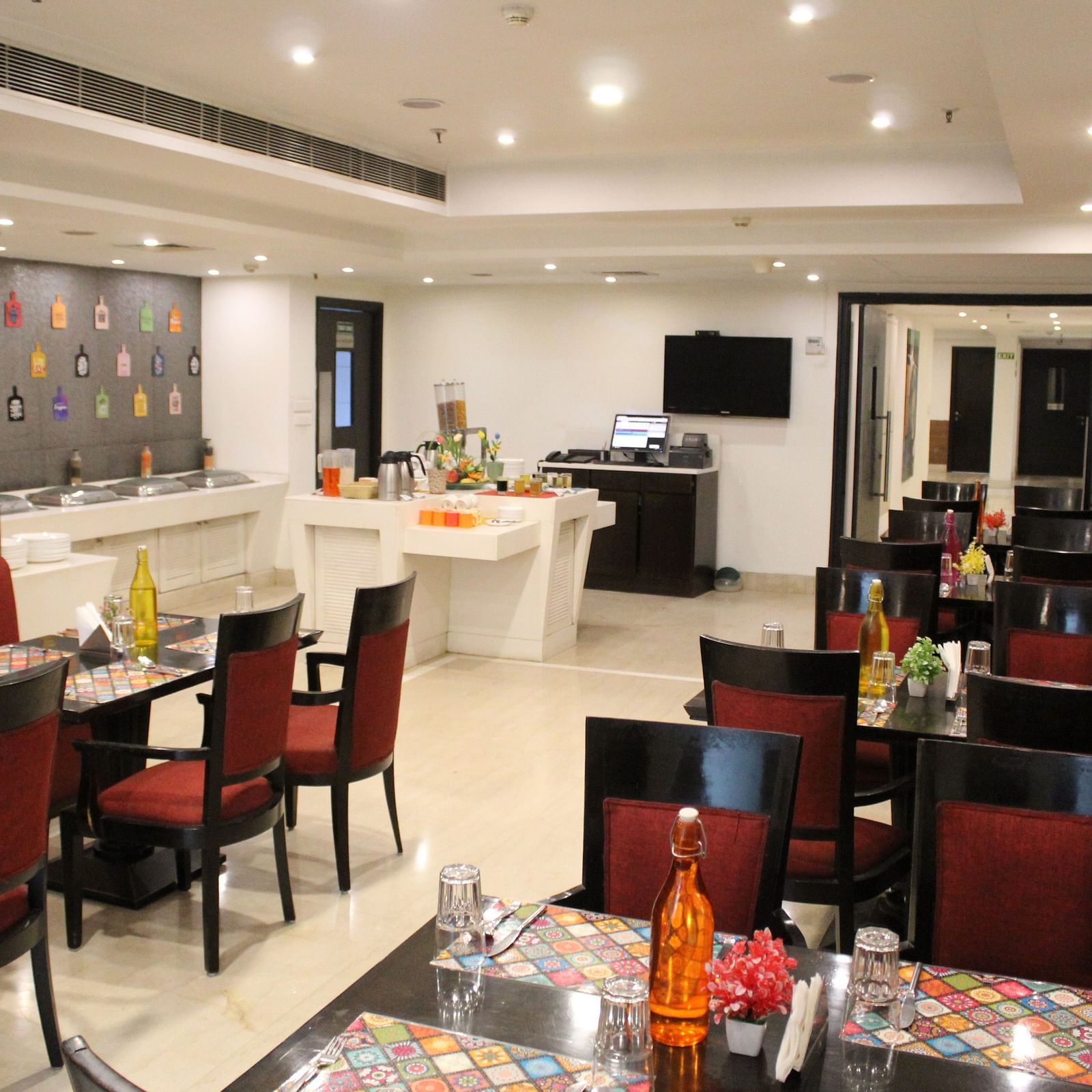 An image of a restaurant several sets of chairs and table where plates, cutlery, napkin, water bottles and other items are kept - Inde Hotels Vista Woods