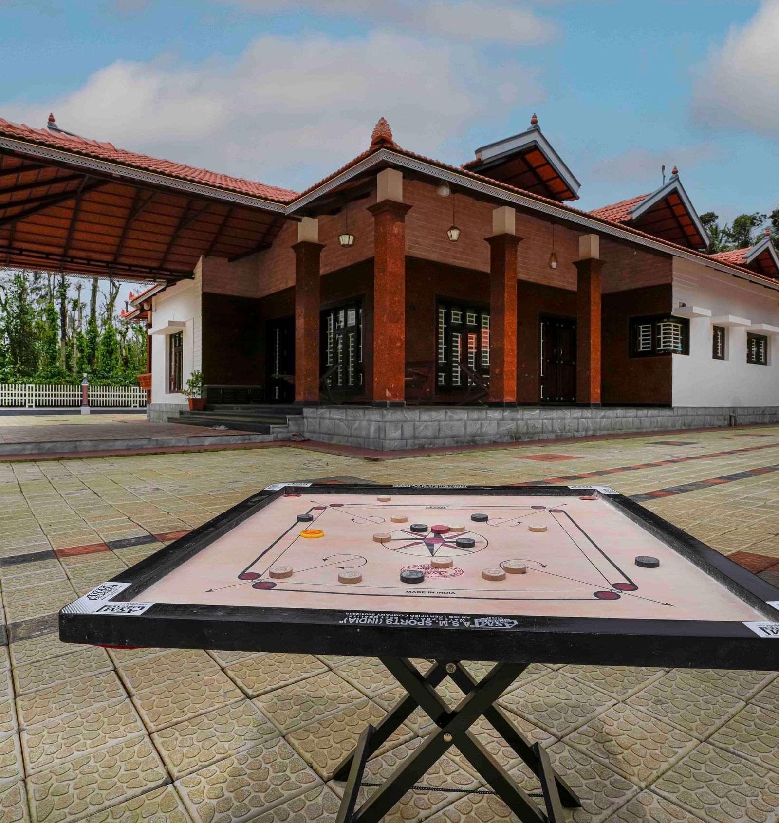 A carrom board is set up outdoors on a paved courtyard, with the traditional architecture of Bhuthanakadu Retreat visible in the background.