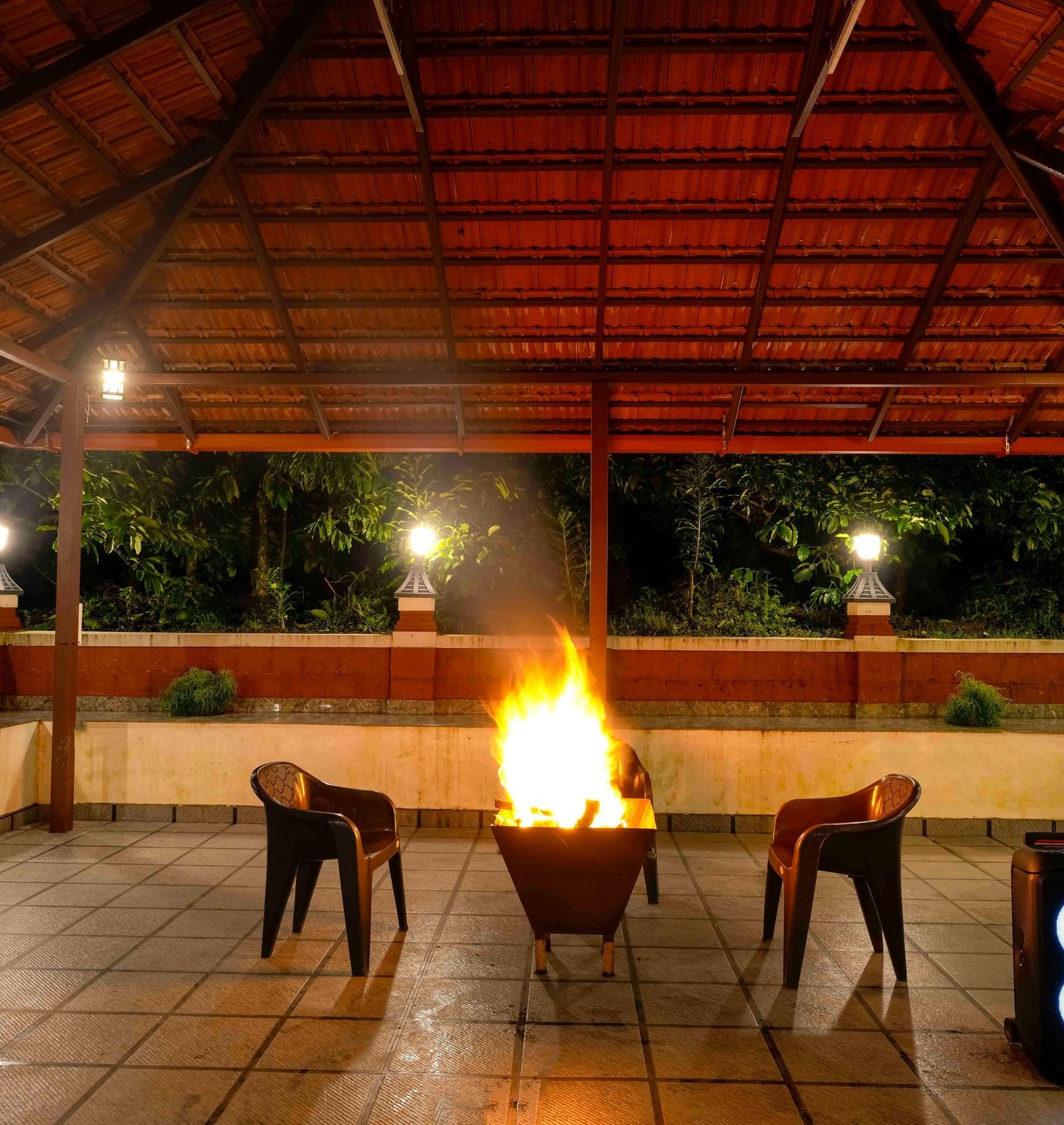 A large bonfire crackles at the center of a covered, open-air pavilion at Bhuthanakadu Retreat, surrounded by chairs under the warm glow of the lights at night.