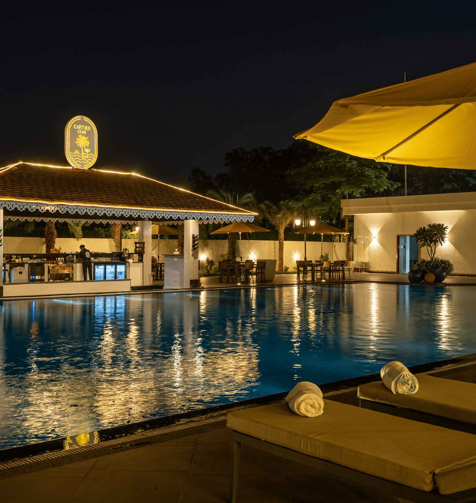 Evening view of the swim-up bar area next to the pool, featuring seating and ambient lighting under an umbrella at Hotel Hukam's Lalit Mahal.