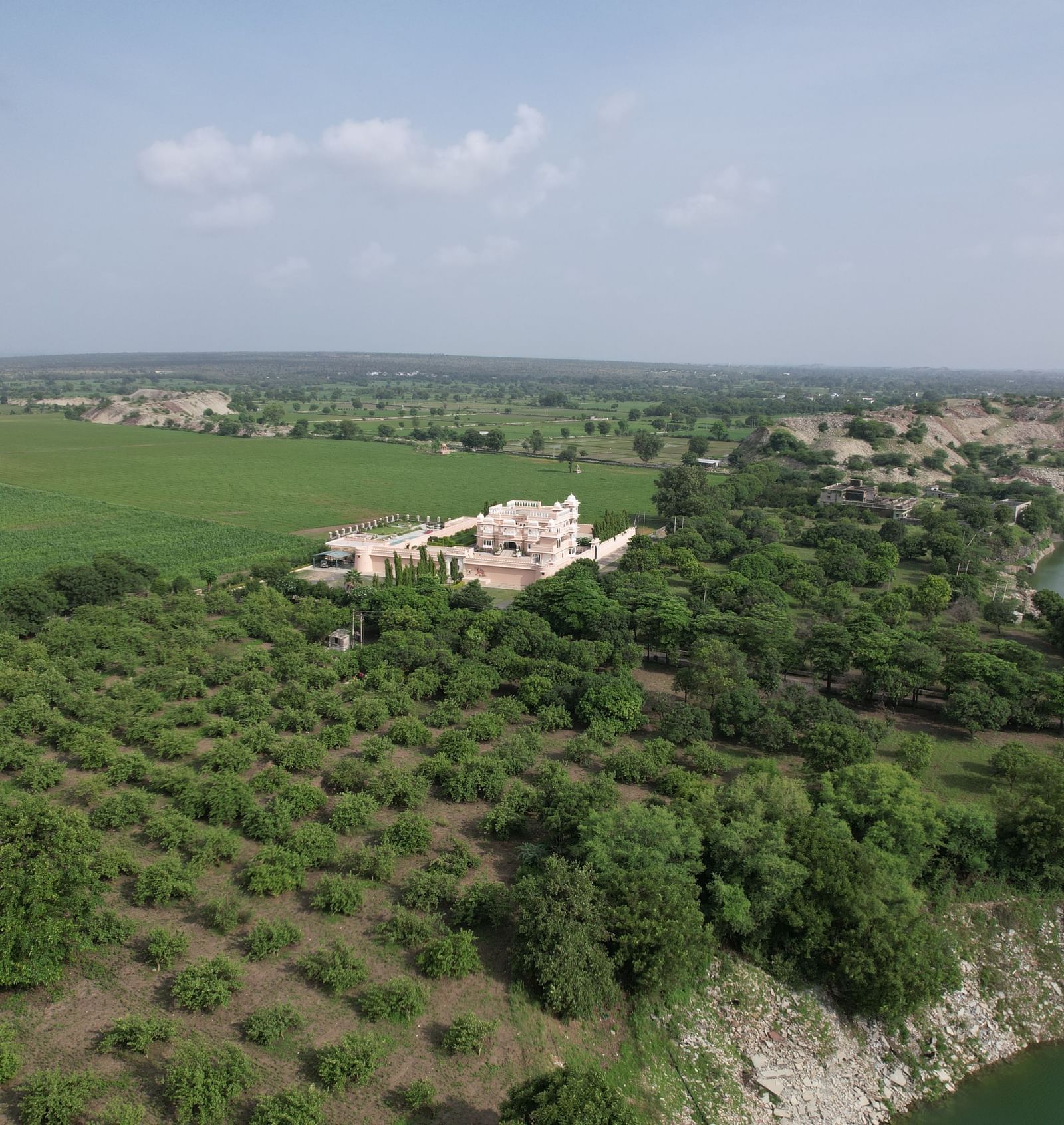 A slightly altered aerial perspective of Mahendra Niwas, emphasizing the sprawling property surrounded by lush greenery and the tranquil lake, with a clearer view of the distant horizon.