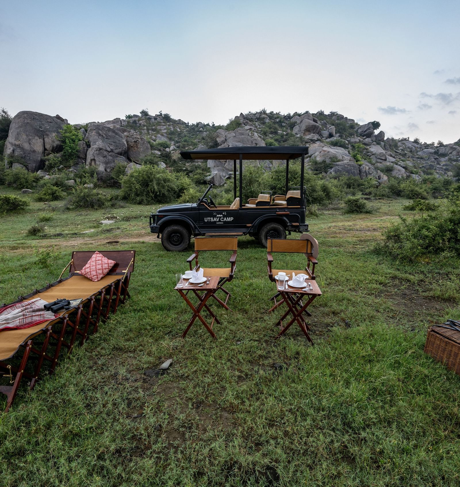 An image of an outdoor seating arrangement with a jeep parked nearby amidst greenery and the backdrop of green mountains - Utsav Camp Sariska