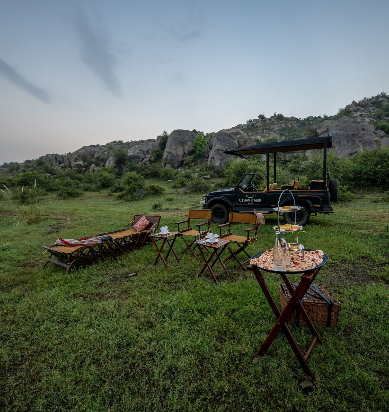 An image of an outdoor seating arrangement with a jeep parked nearby amidst greenery and the backdrop of green mountains - Utsav Camp Sariska