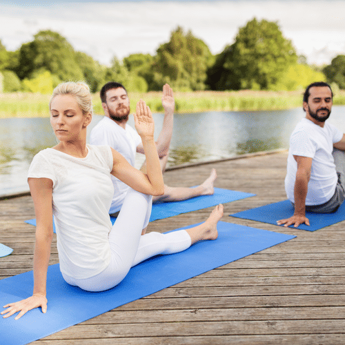 Group of people practicing seated spinal twist yoga poses on mats by a lakeside wooden deck, surrounded by greenery and calm water