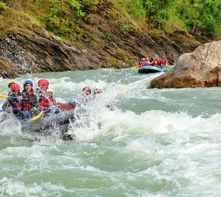alt-text A group of people wearing red life jackets and helmets rafting down a fast-moving river surrounded by lush, green hills.